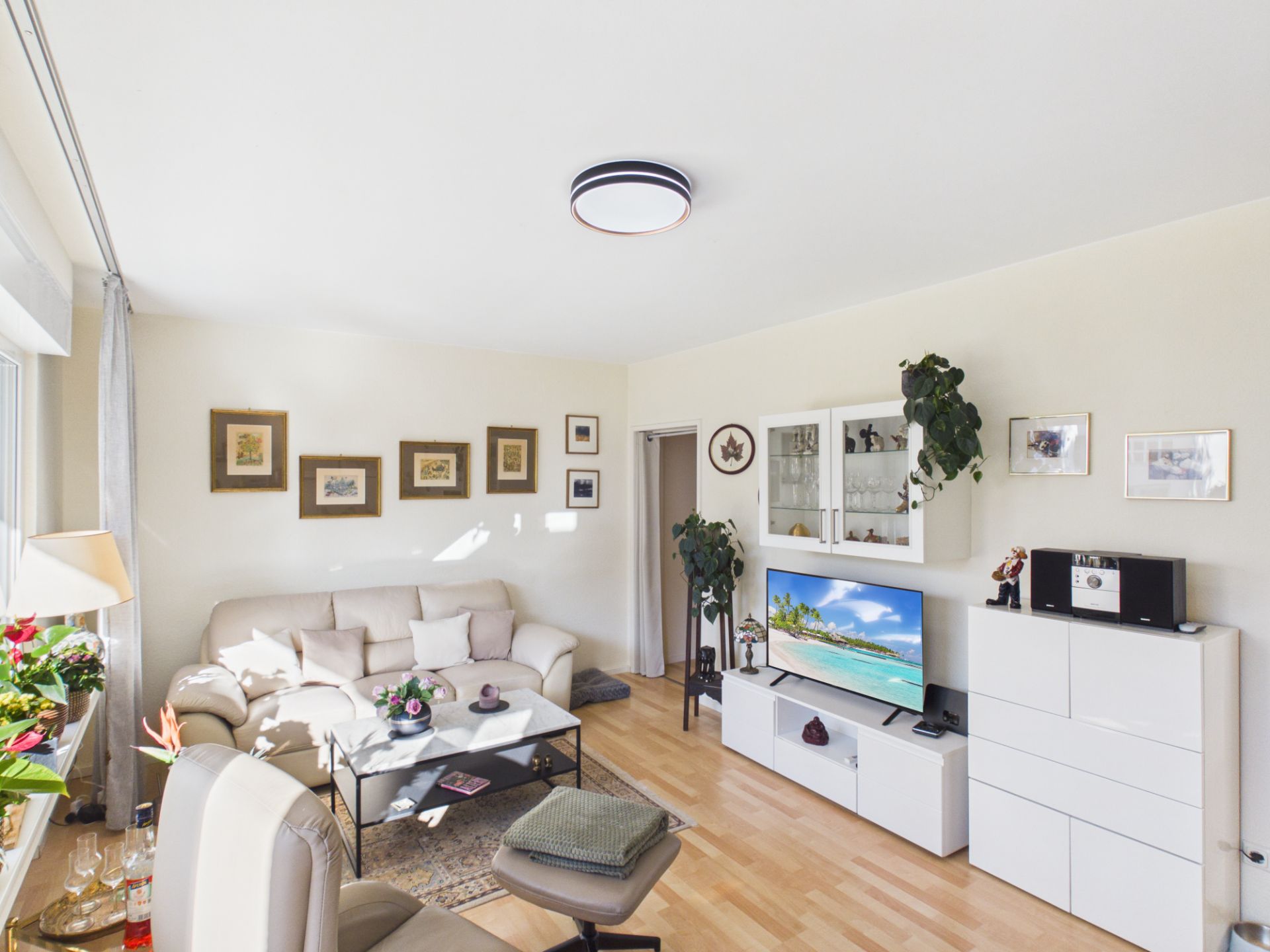Helles Wohnzimmer mit Sofa, Fernseher, Bildern an der Wand und Holzboden.