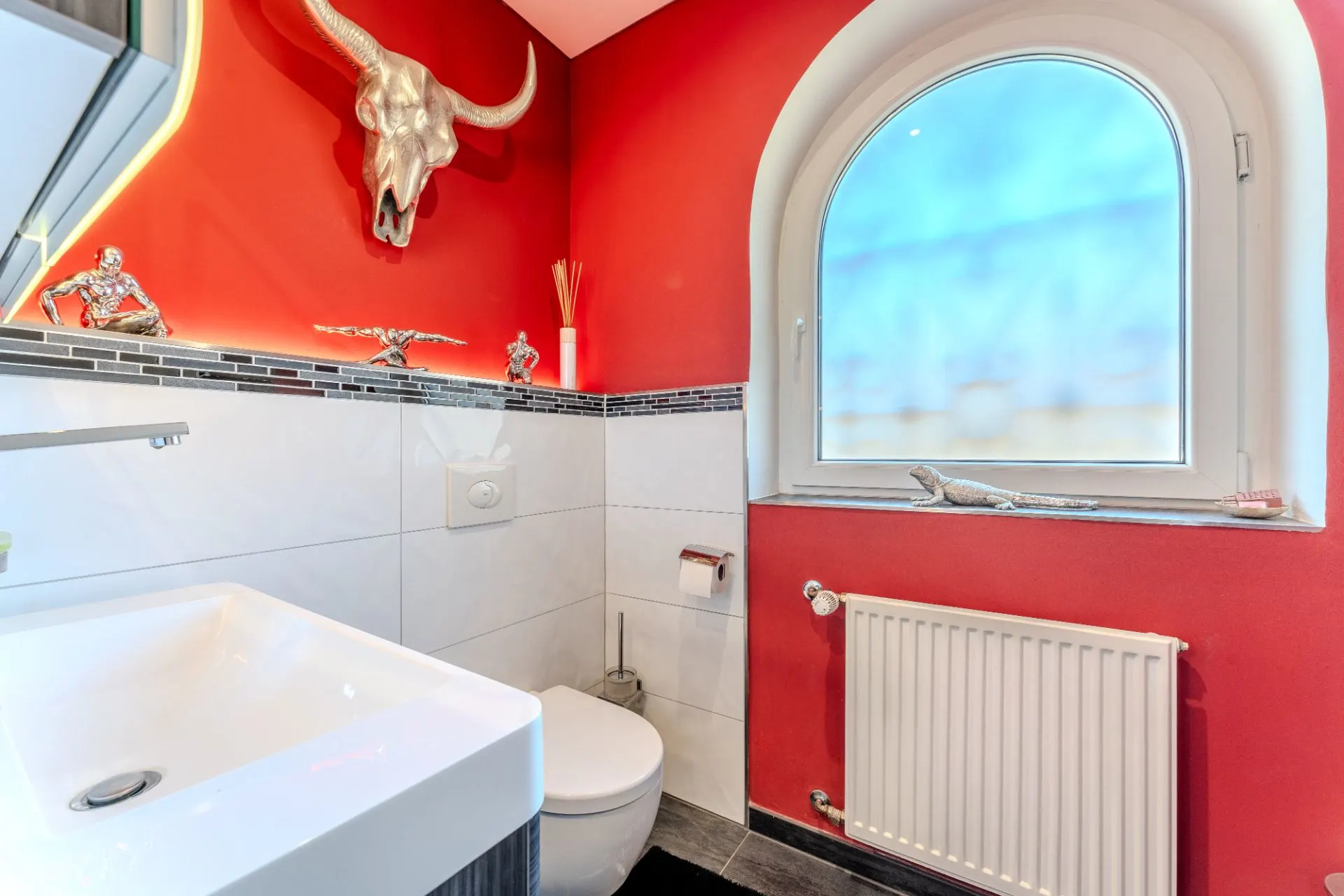 Kleines Badezimmer mit roter Wand, Waschbecken, Toilette und großem Fenster.