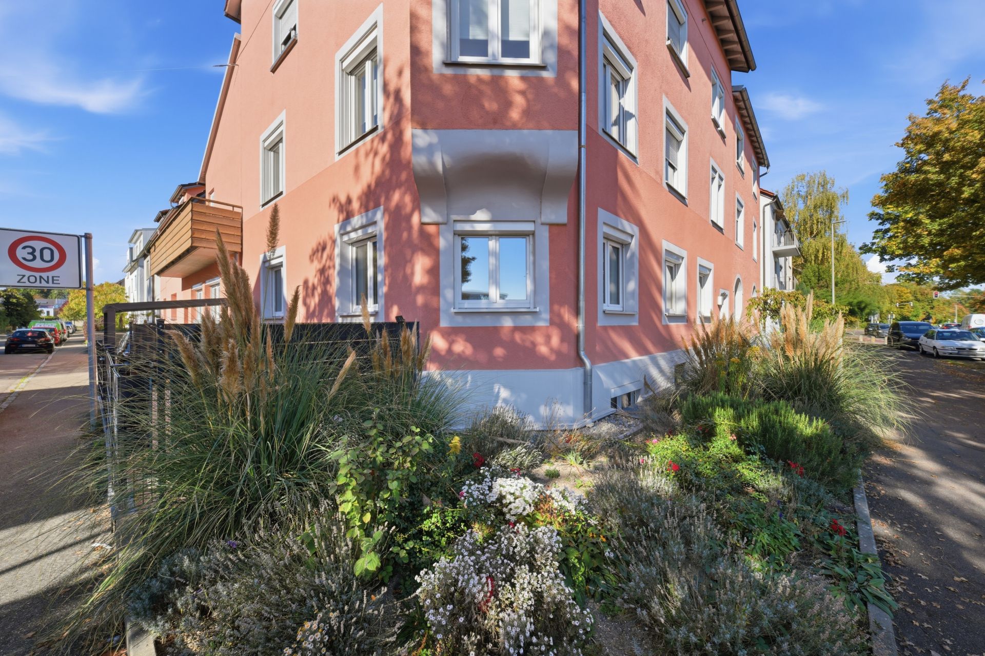 Eckhaus mit rosafarbener Fassade, weißen Fenstern und einem Vorgarten mit Sträuchern und Blumen.