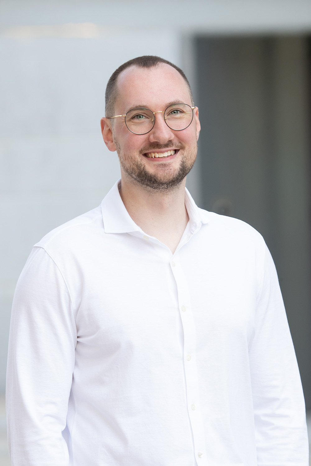 Ein lächelnder Mann mit Brille und weißem Hemd steht vor einem unscharfen Hintergrund.