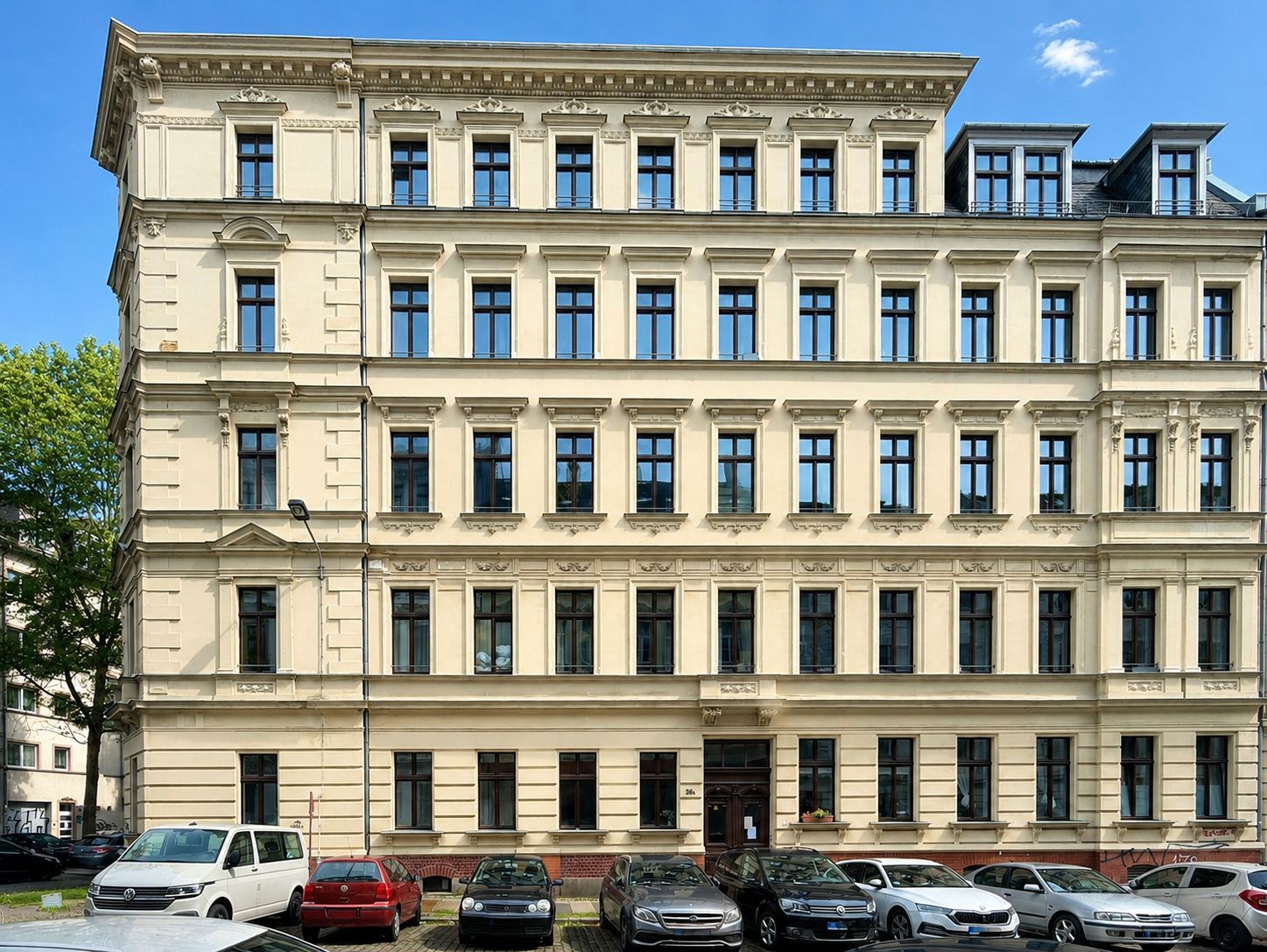 Vierstöckiges Wohngebäude mit vielen Fenstern und parkenden Autos davor.