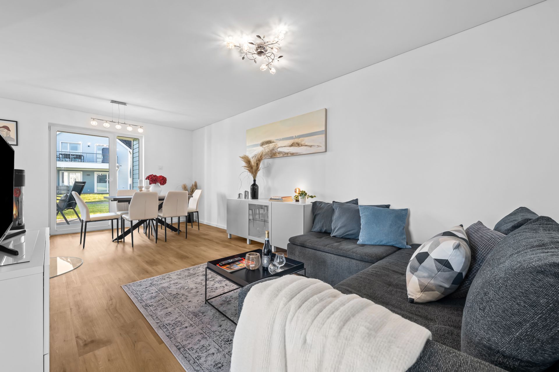 Modernes Wohnzimmer mit grauem Sofa, Esstisch, großer Fensterfront und Holzboden.