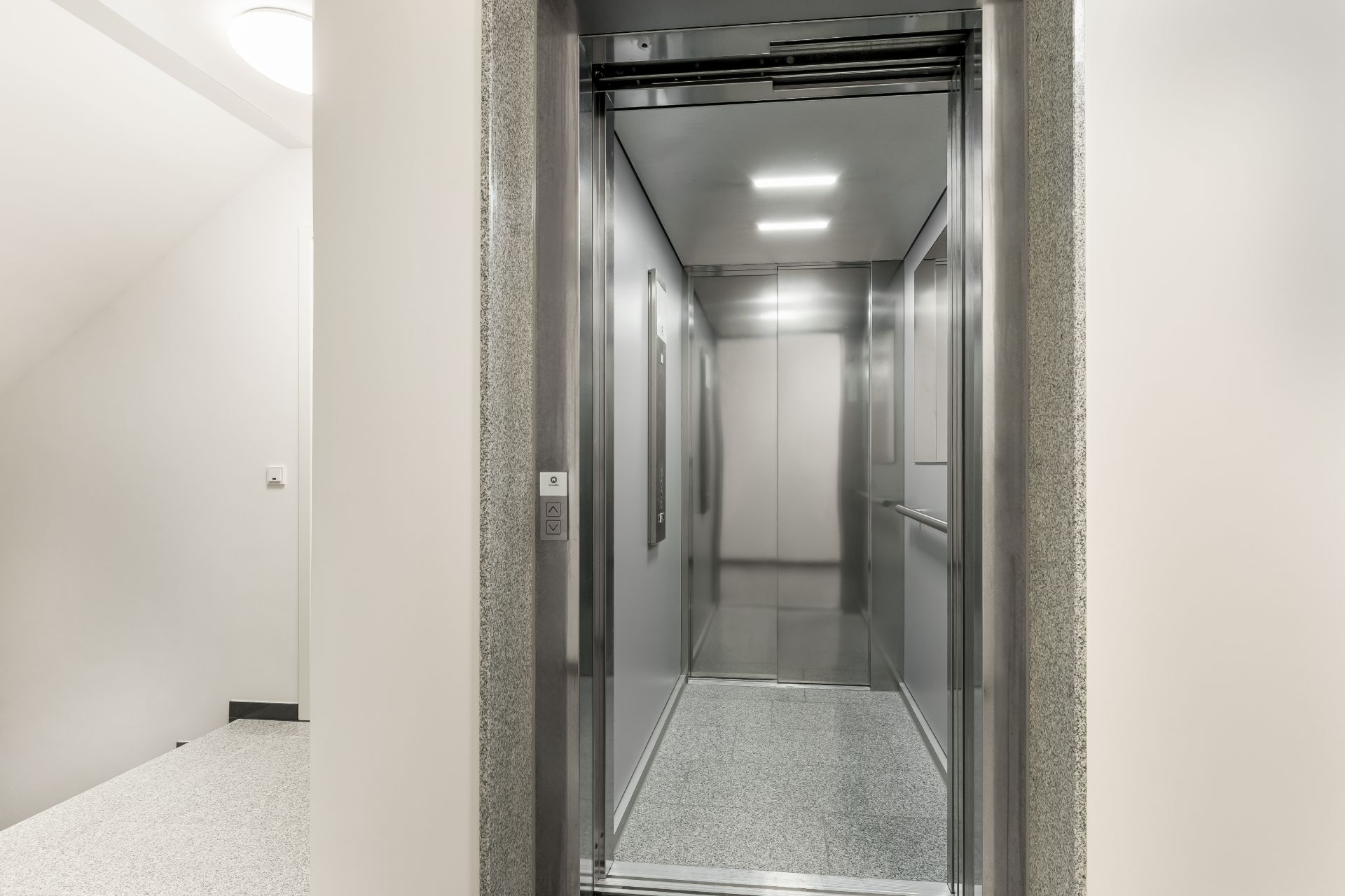 Offener Aufzug mit silbernem Innenraum und beleuchtetem Deckenelement neben einer Treppe.
