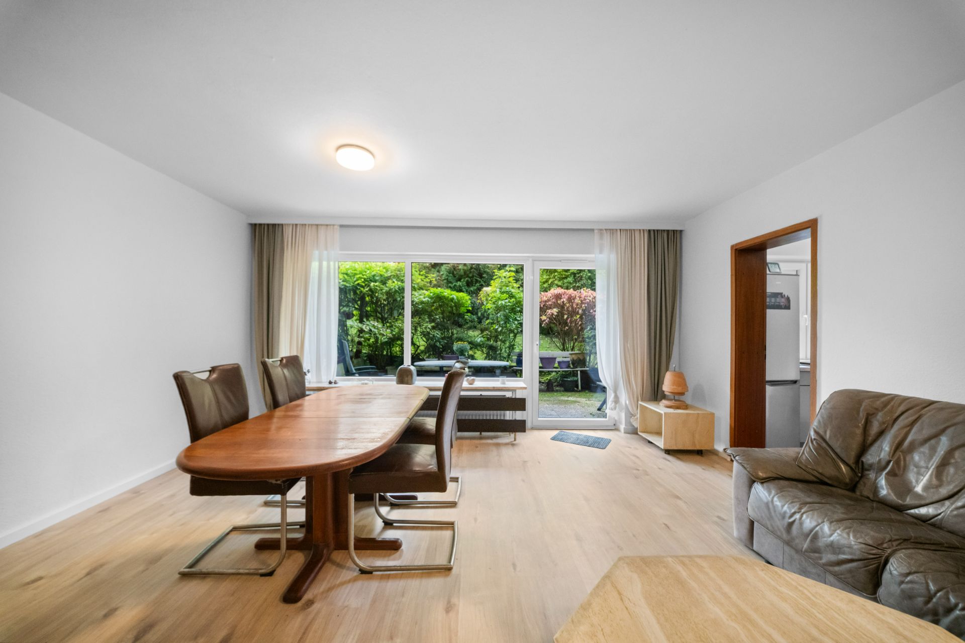 Helles Wohnzimmer mit Esstisch, Stühlen, Sofa und Blick auf den Garten durch große Fenster.