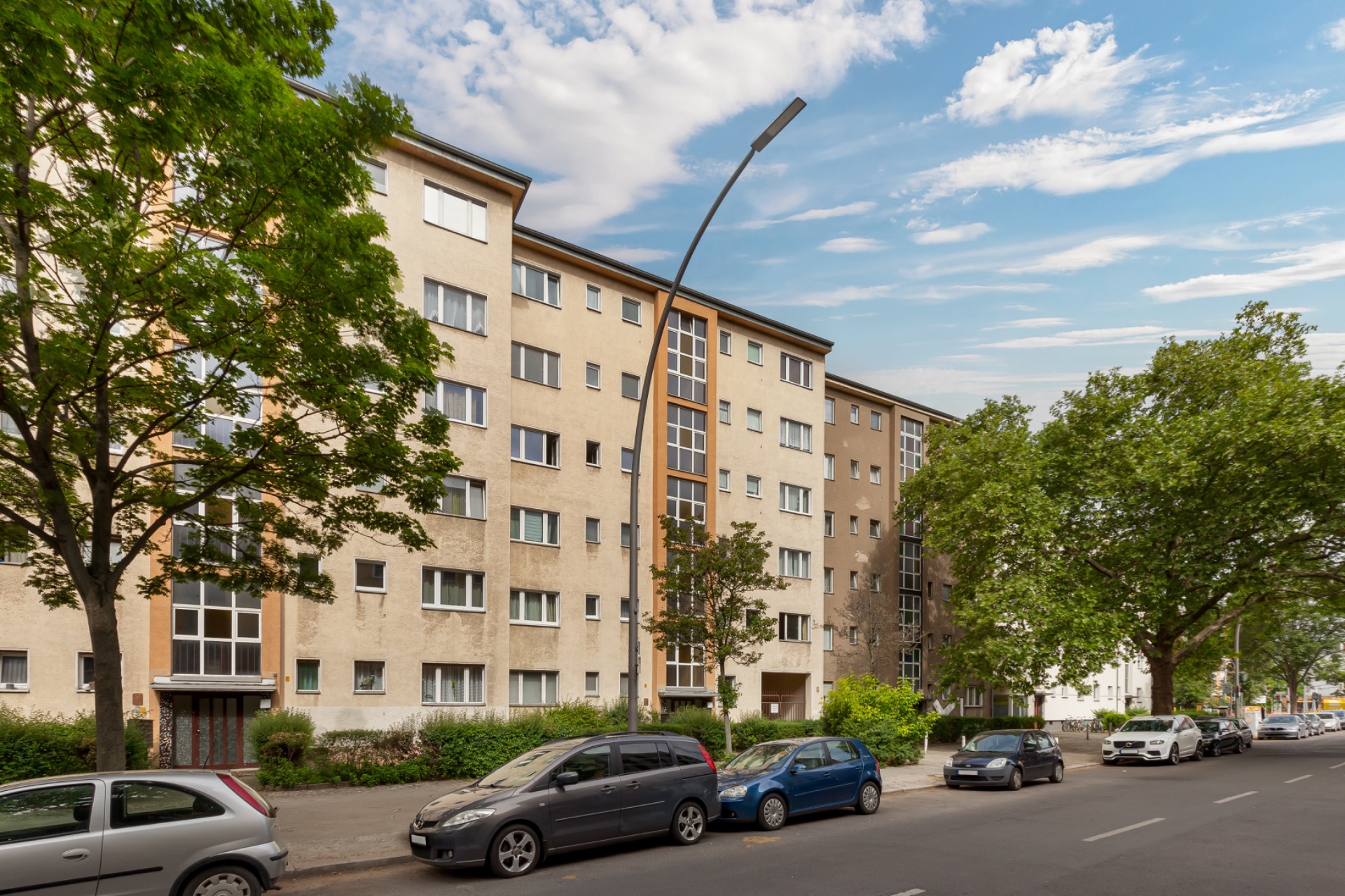 Mehrstöckiges Wohnhaus mit mehreren Eingängen, Bäumen und geparkten Autos an der Straße.