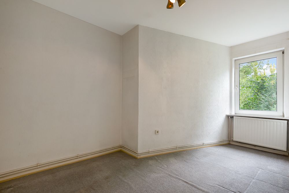 Leeres Zimmer mit Teppichboden, weißer Wand und Fenster mit Blick ins Grüne.