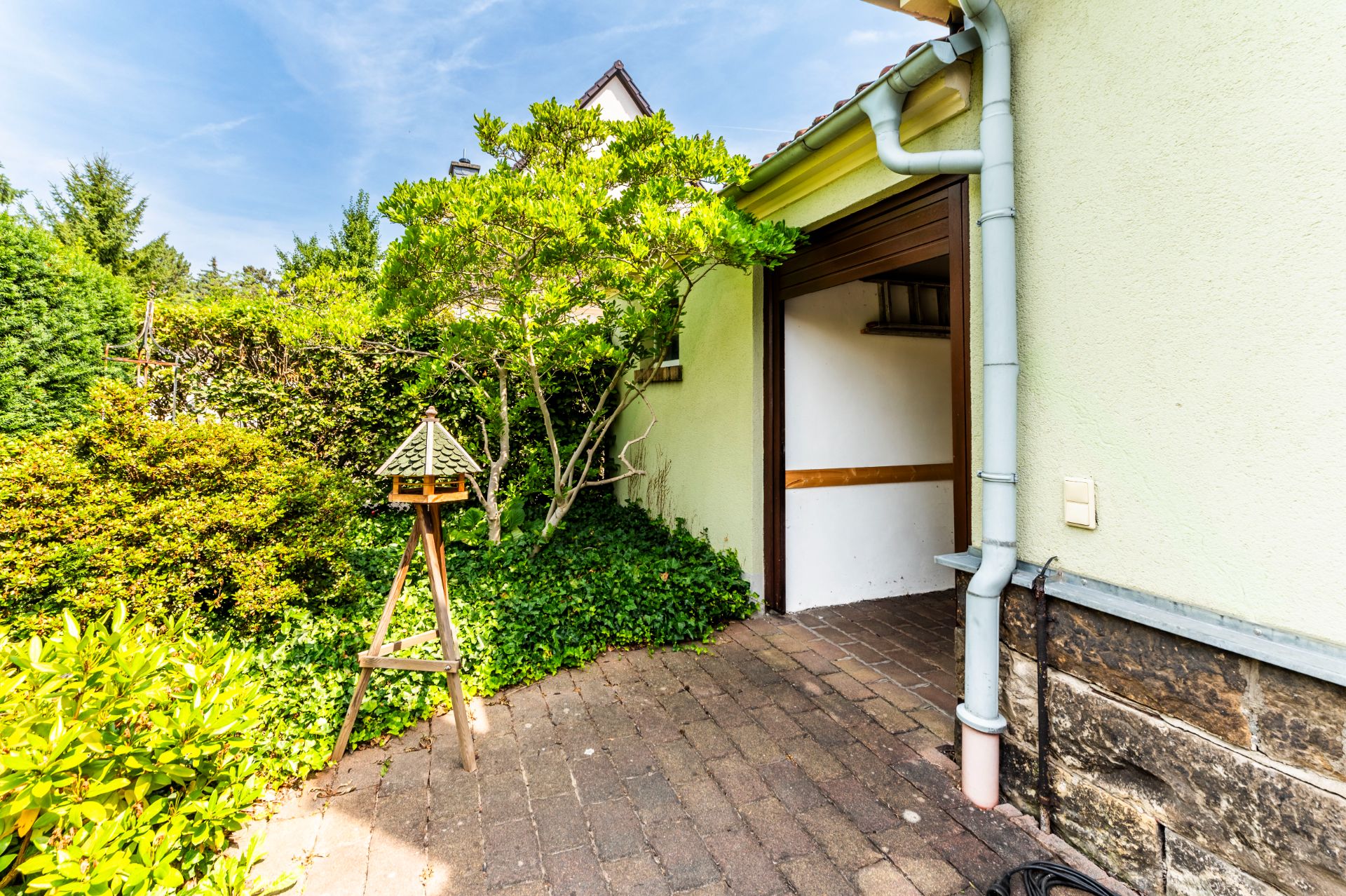Gepflasterter Hauseingang mit offener Tür, Vogelhaus und grünen Sträuchern im Garten.