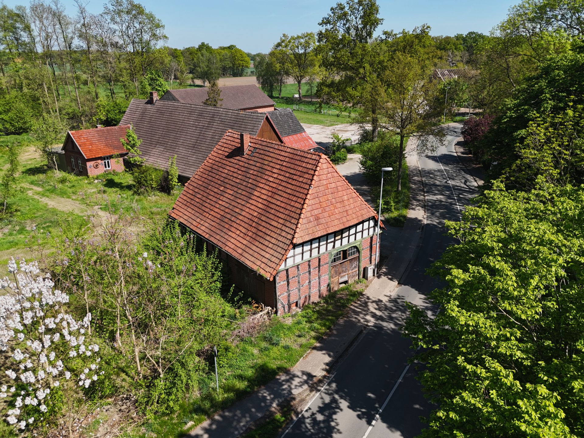 Zwei Backsteingebäude mit roten Dächern an einer Landstraße, umgeben von Bäumen und Wiese.