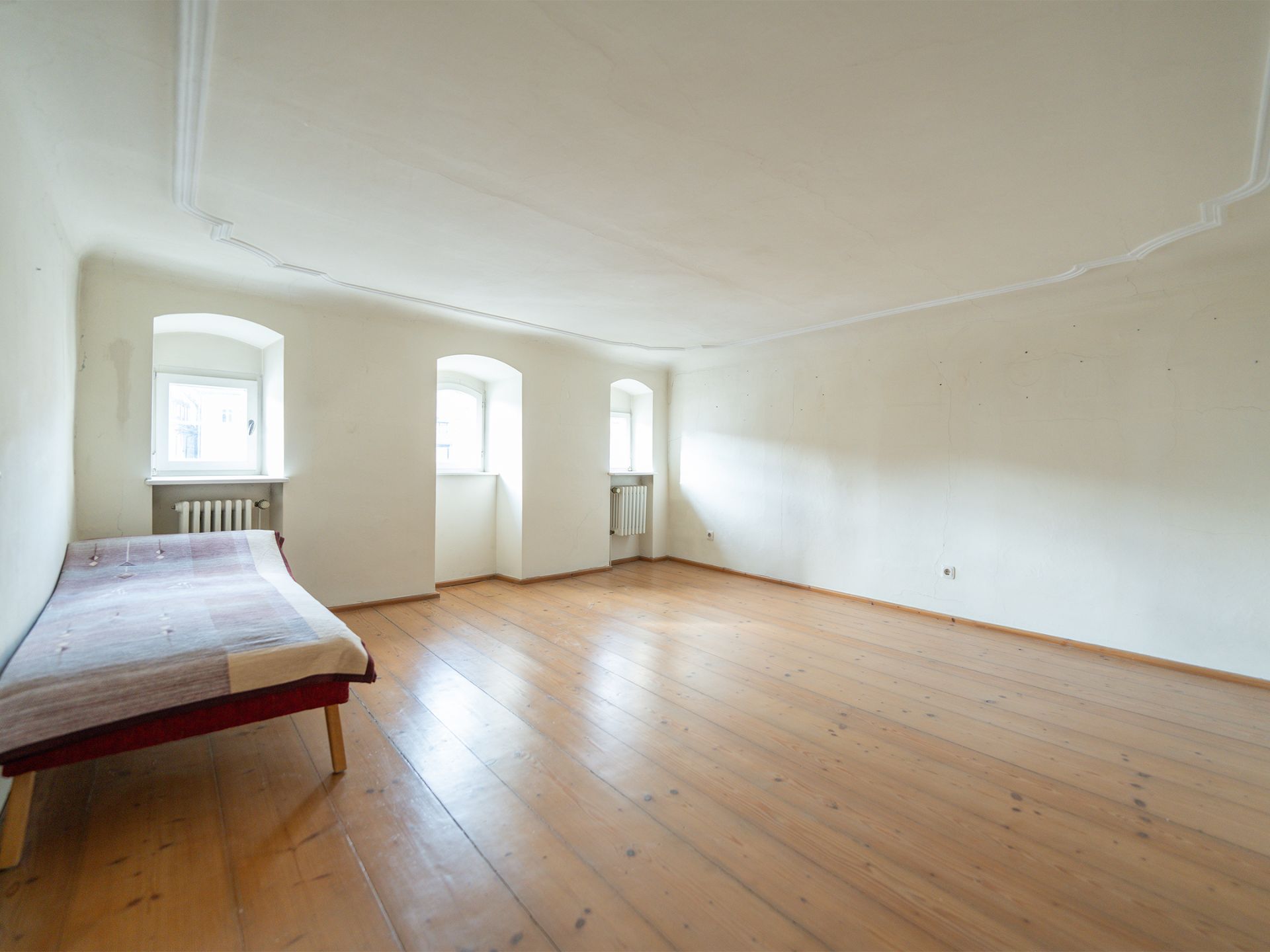 Leerer Raum mit Holzboden, drei Fenstern und einem einfachen Bett links.