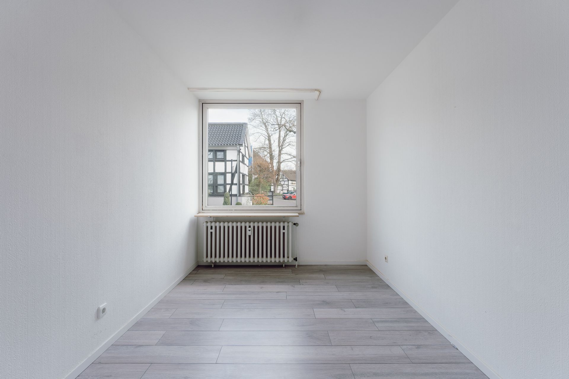 Leerer Raum mit weiß gestrichenen Wänden, Fenster, Heizkörper und hellem Holzfußboden.