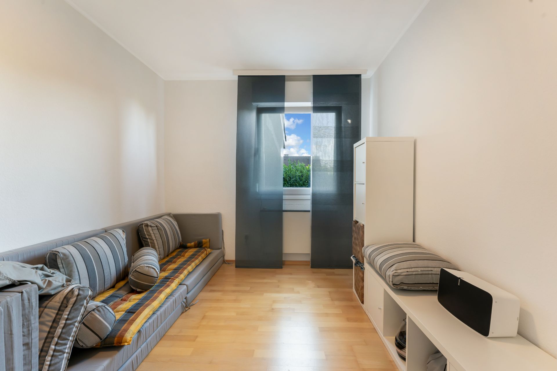Helles Wohnzimmer mit Sofa, Regal und Fenster mit dunklen Vorhängen.