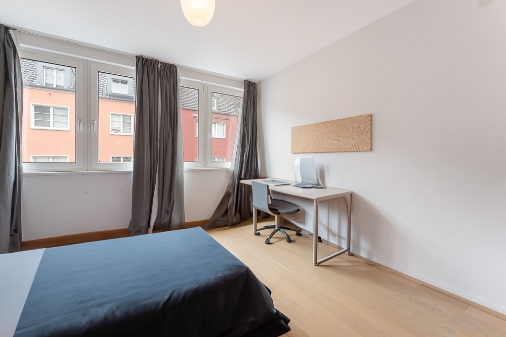 Modernes Schlafzimmer mit Schreibtisch, Bett und großen Fenstern mit grauen Vorhängen.