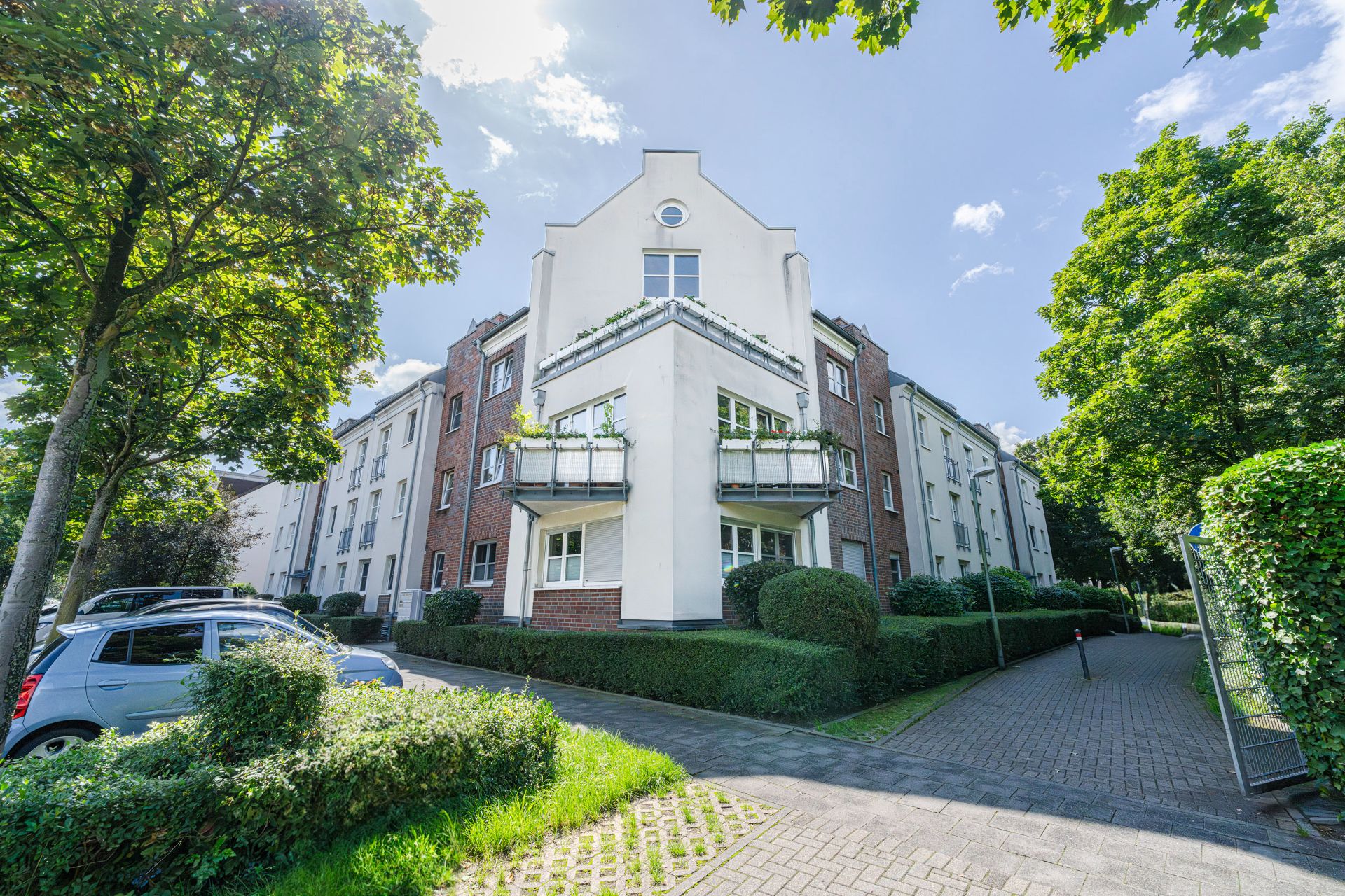 Mehrstöckiges Wohnhaus mit Balkonen, umgeben von Bäumen und parkenden Autos.