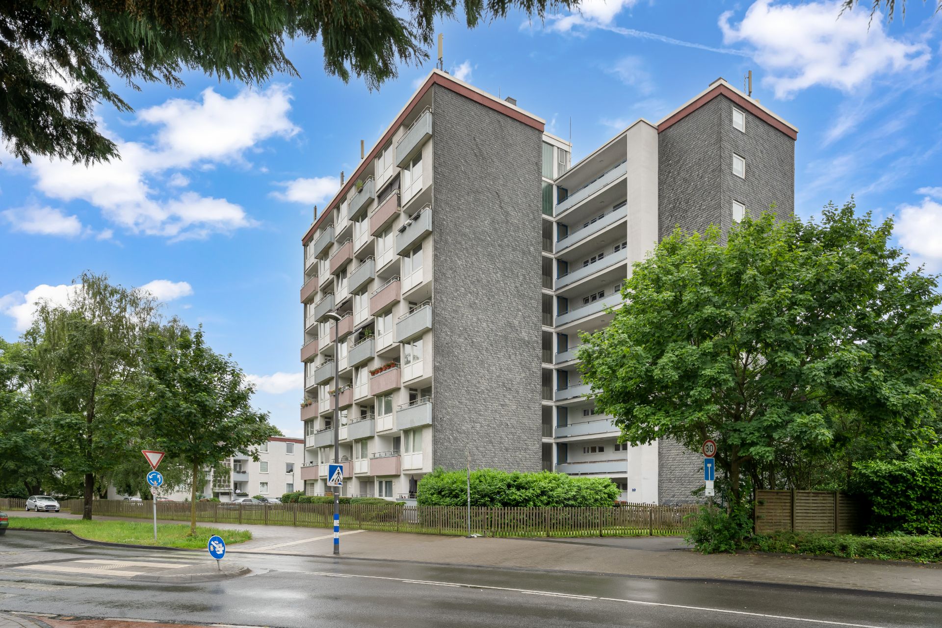 Mehrstöckiges Wohnhaus mit Balkonen, umgeben von Bäumen und einer Straße im Vordergrund.