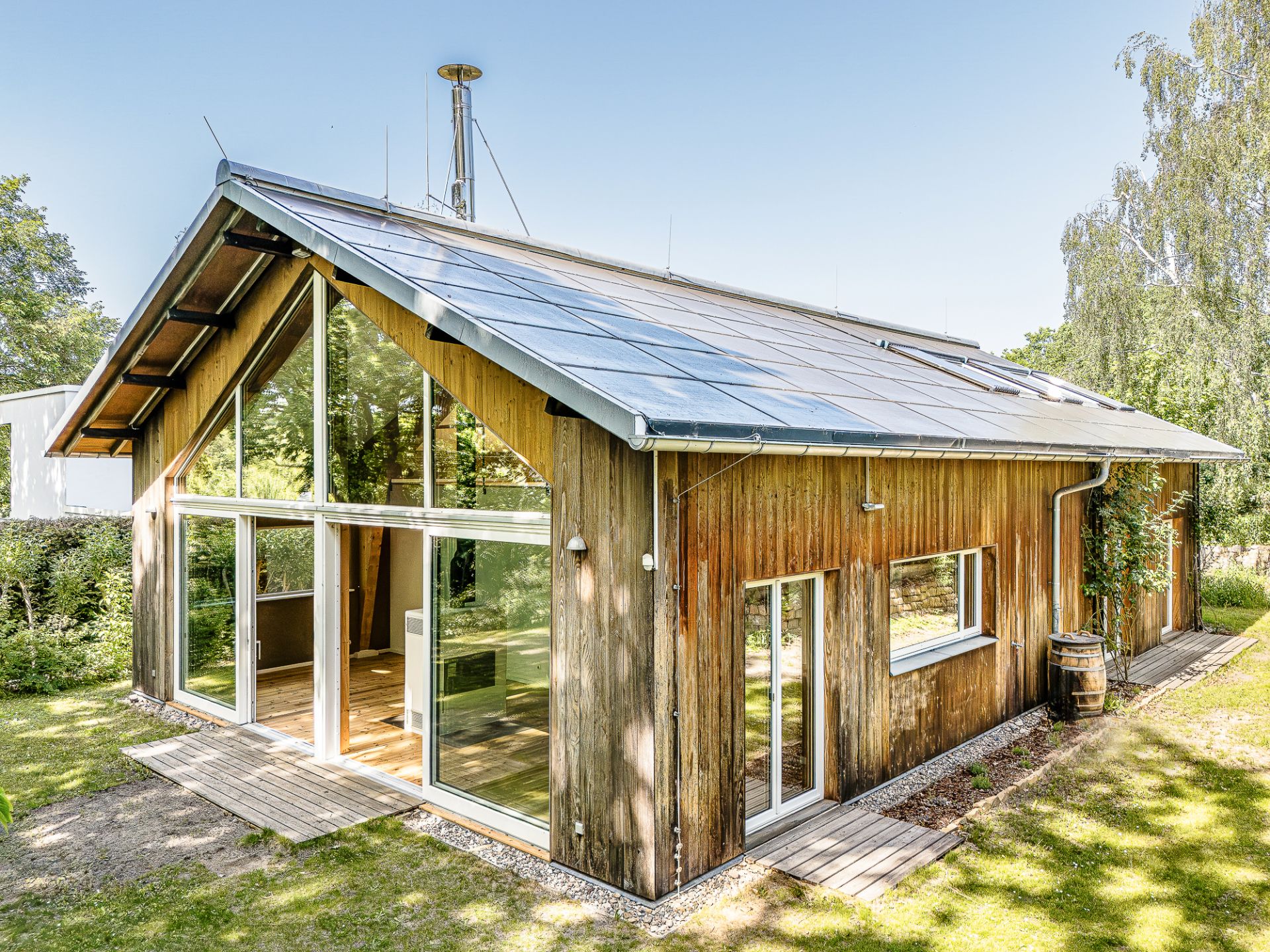 Modernes Holzhaus mit großen Fenstern und Solarpaneelen auf dem Dach, umgeben von Grün.