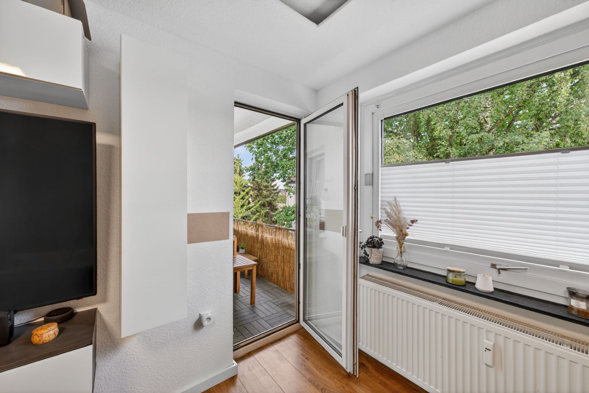 Wohnzimmer mit offenem Zugang zum Balkon und großem Fenster mit Blick auf Bäume.