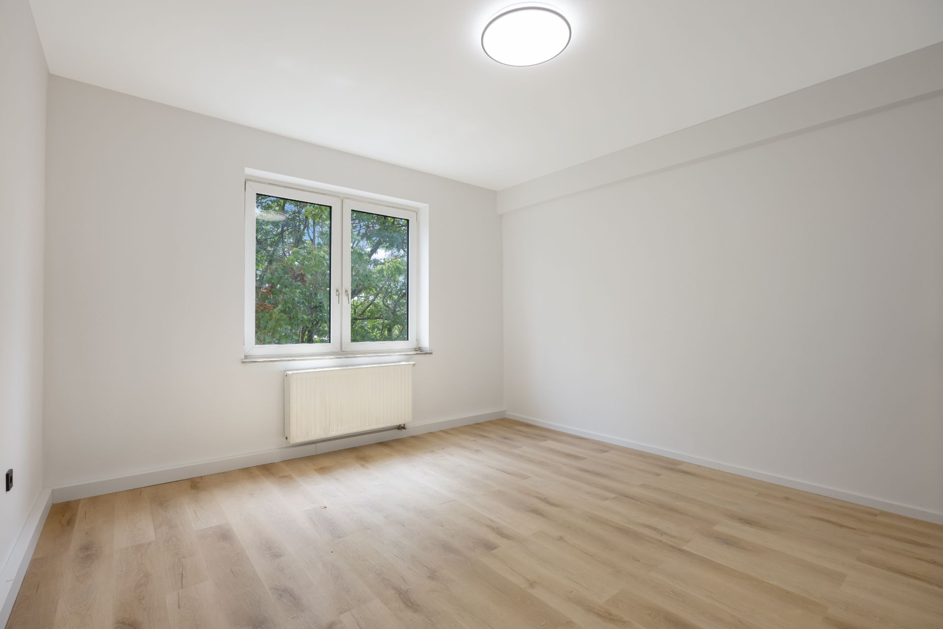Leerer Raum mit hellem Holzboden, weißer Wand, Fenster und Heizkörper.