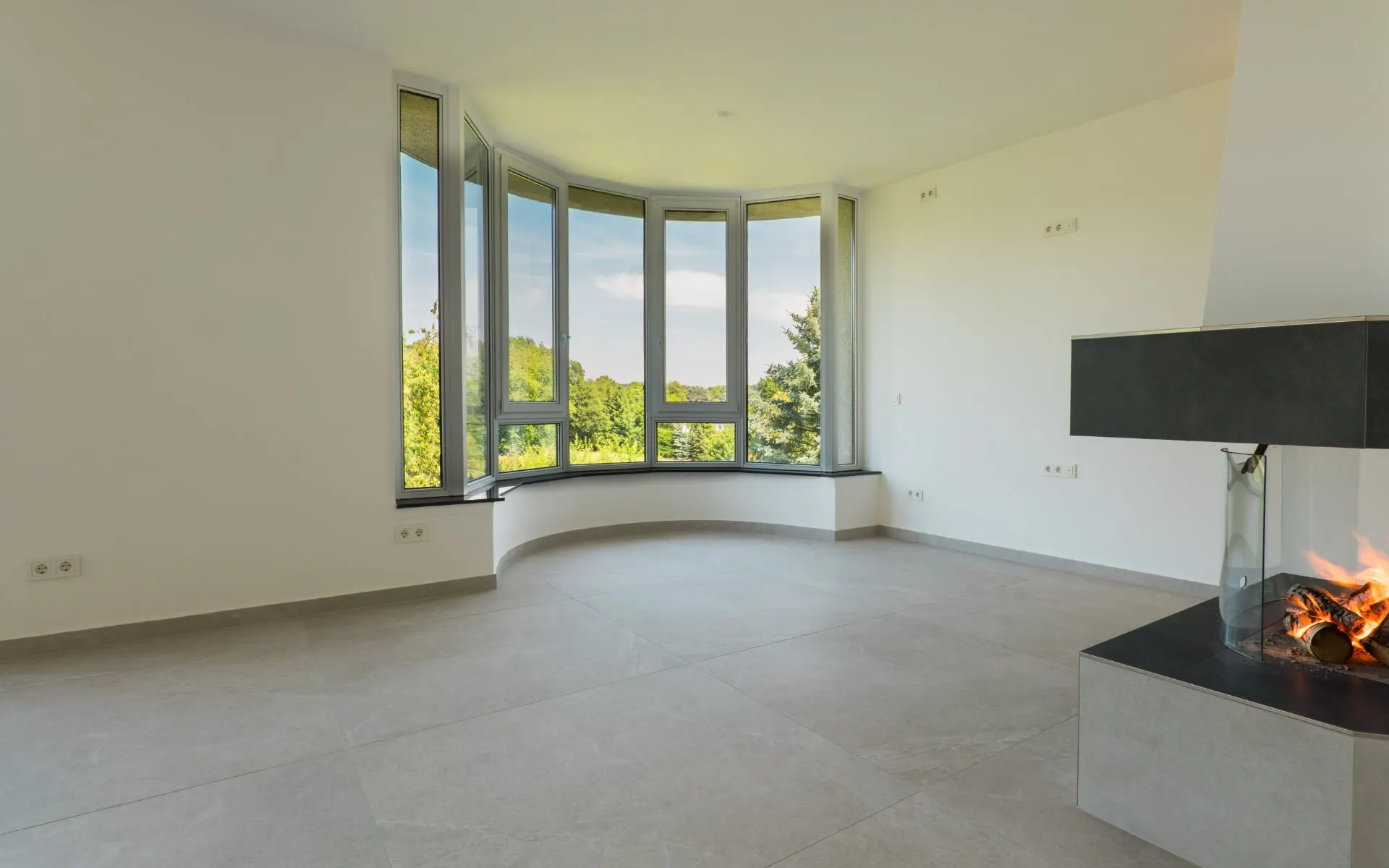 Helles Wohnzimmer mit großen Fenstern und modernem Kamin, Blick auf grüne Bäume.