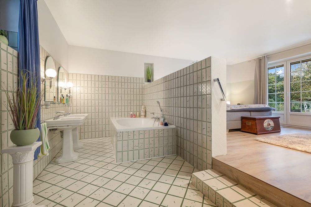 Offenes Badezimmer mit gefliester Badewanne und Waschbecken, angrenzend an einen Wohnbereich mit Sofa.