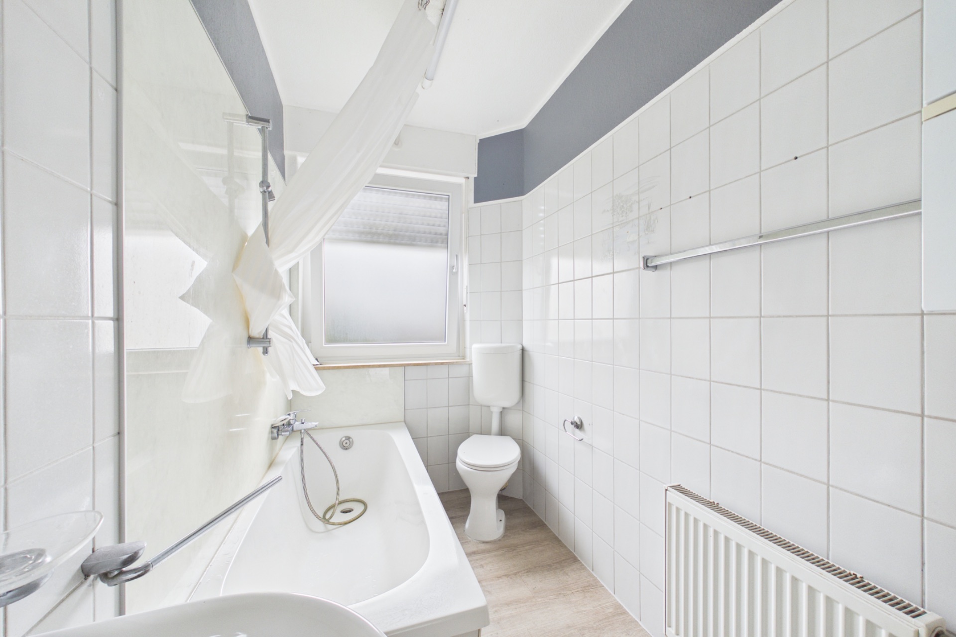 Helles Badezimmer mit Badewanne, Toilette, Waschbecken, Handtuchheizkörper und Fenster.