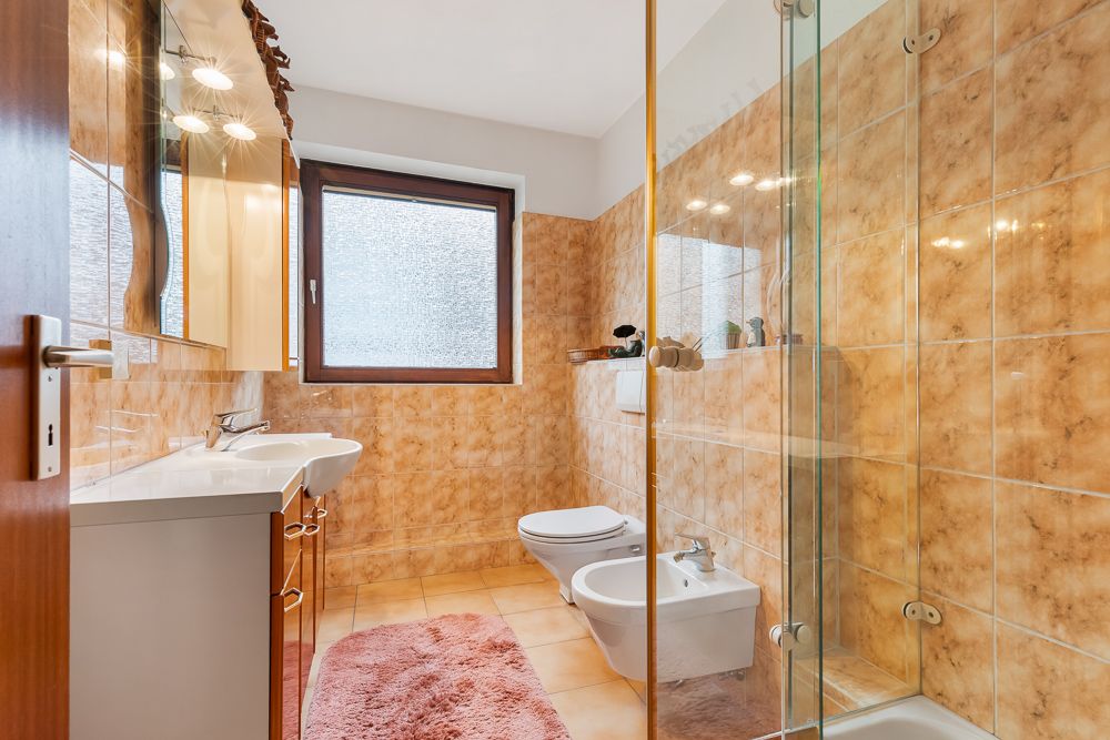 Badezimmer mit braunen Fliesen, Dusche, Bidet, WC, Doppelwaschbecken und Fenster.