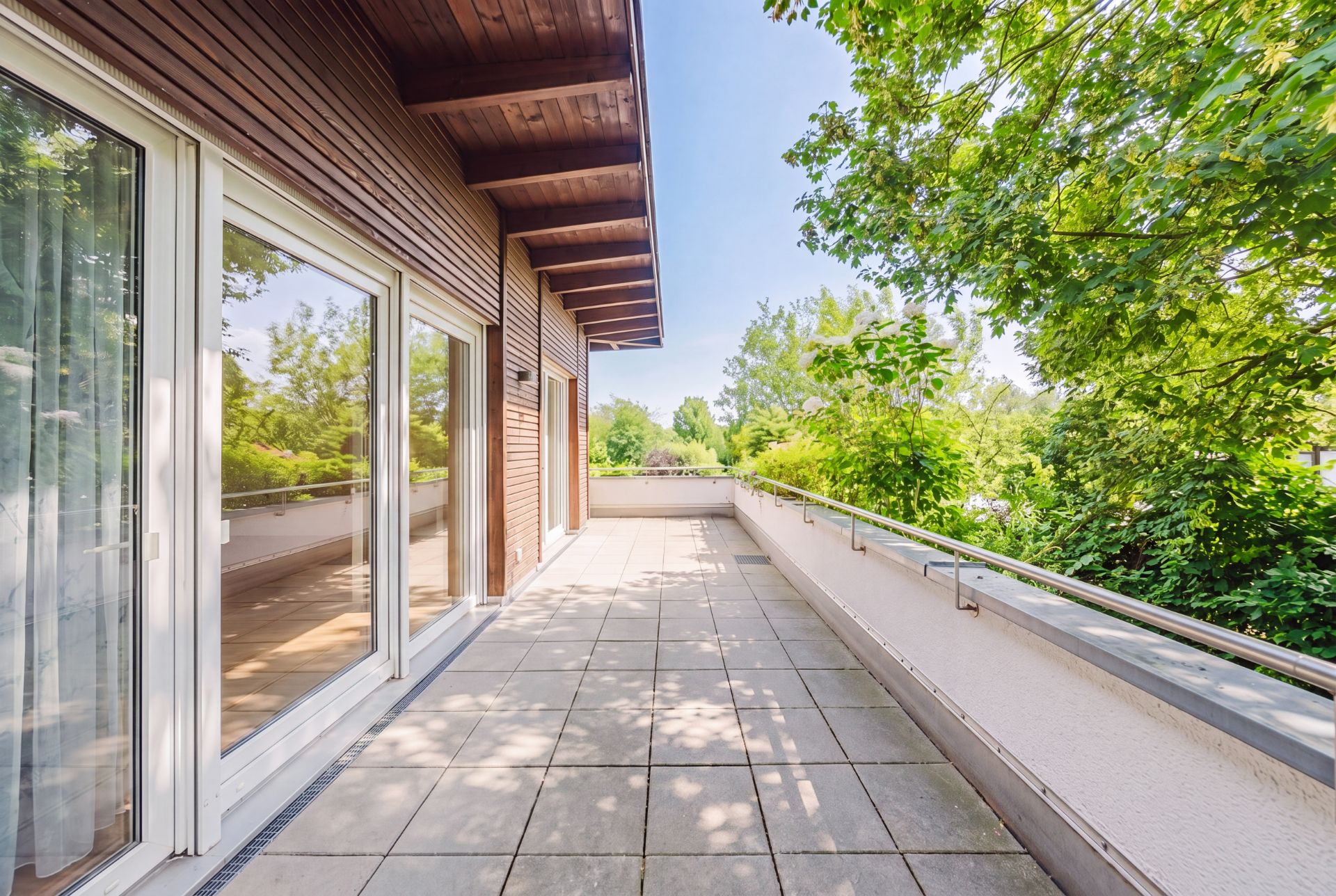 Große Terrasse mit Fliesenboden, Glasgeländer und Blick ins Grüne, angrenzend an ein Wohnhaus.