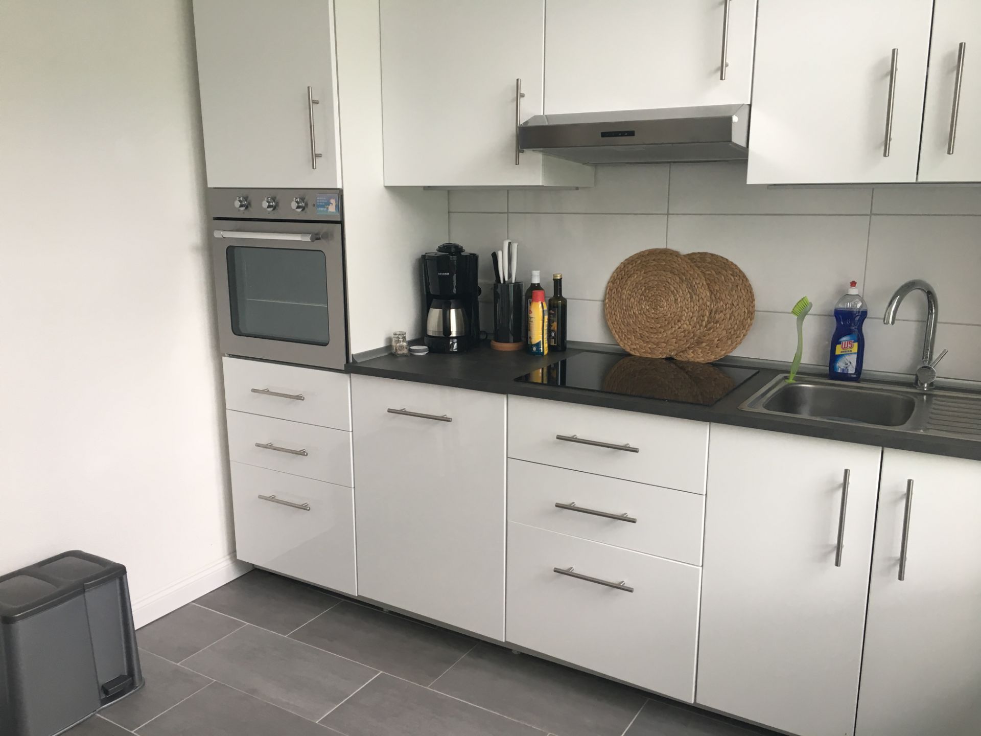 Moderne weiße Küche mit Backofen, Kochfeld, Kaffeemaschine und Spüle.