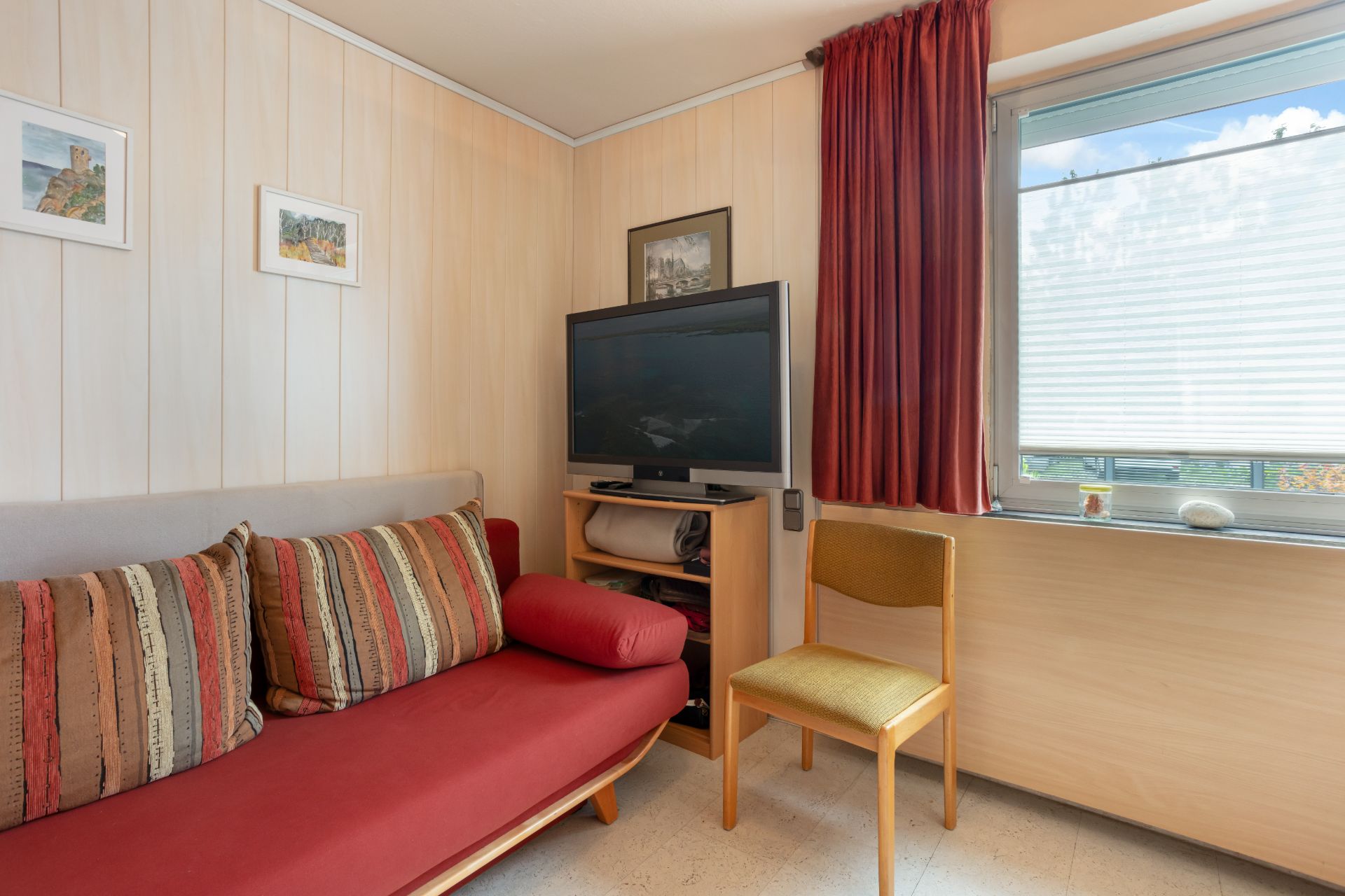 Wohnzimmer mit rotem Sofa, gestreiften Kissen, Fernseher, Stuhl und Fenster mit Vorhang.