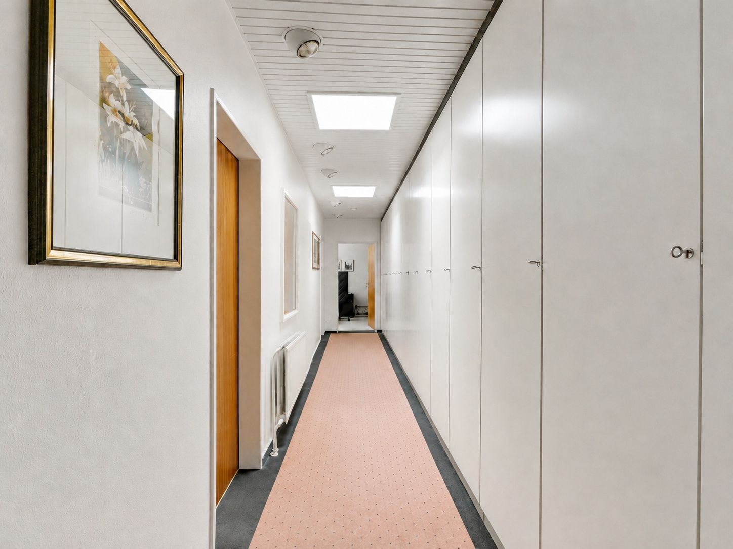 Langer Flur mit weißen Einbauschränken, Bildern an der Wand und hellem Teppichläufer.