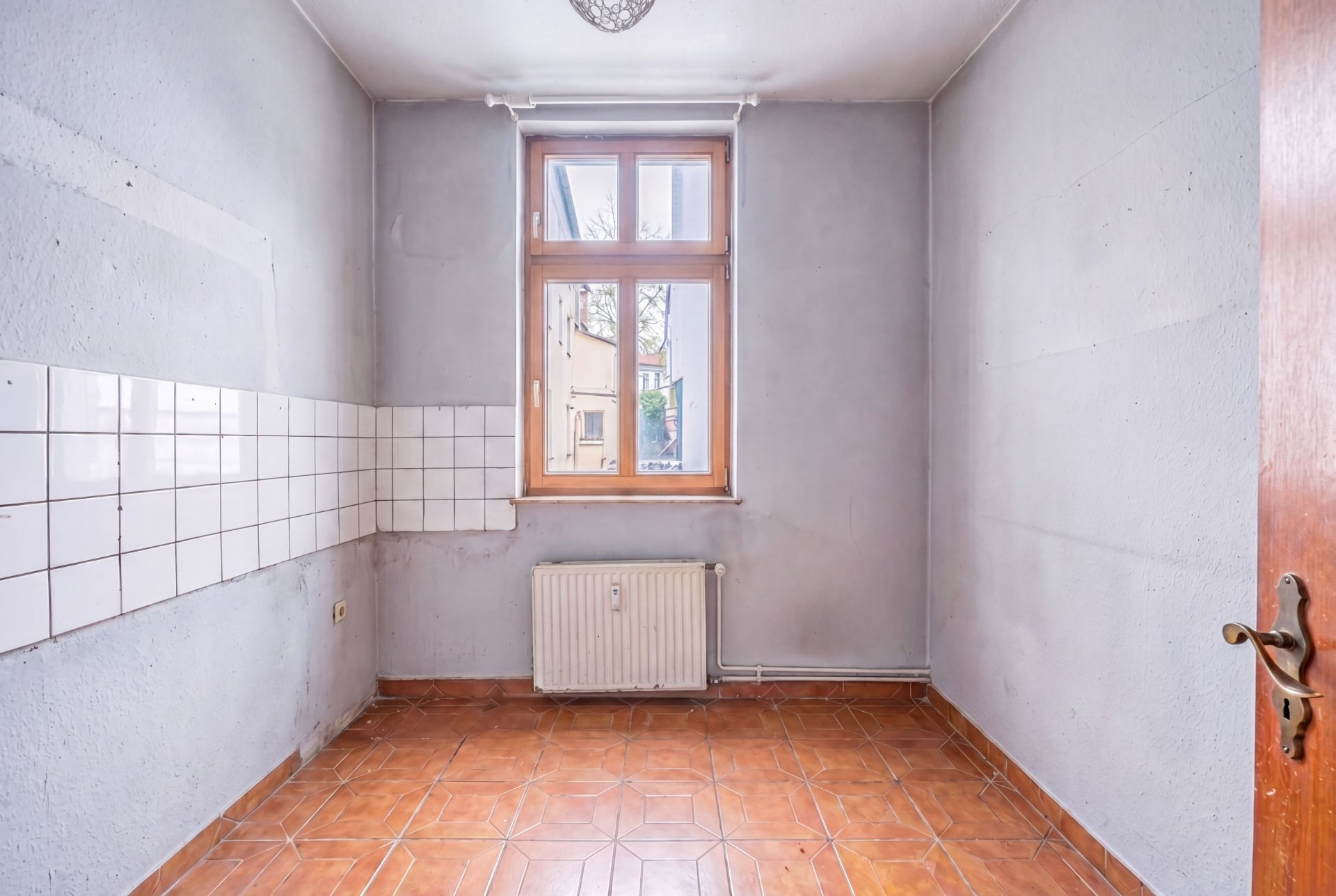 Leerer Raum mit braunen Fliesen, weißem Fliesenspiegel und Fenster mit Heizkörper darunter.