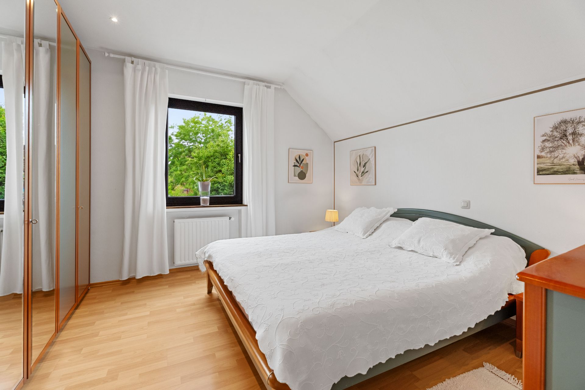 Helles Schlafzimmer mit Doppelbett, Holzschrank, Fenster und weißen Vorhängen.