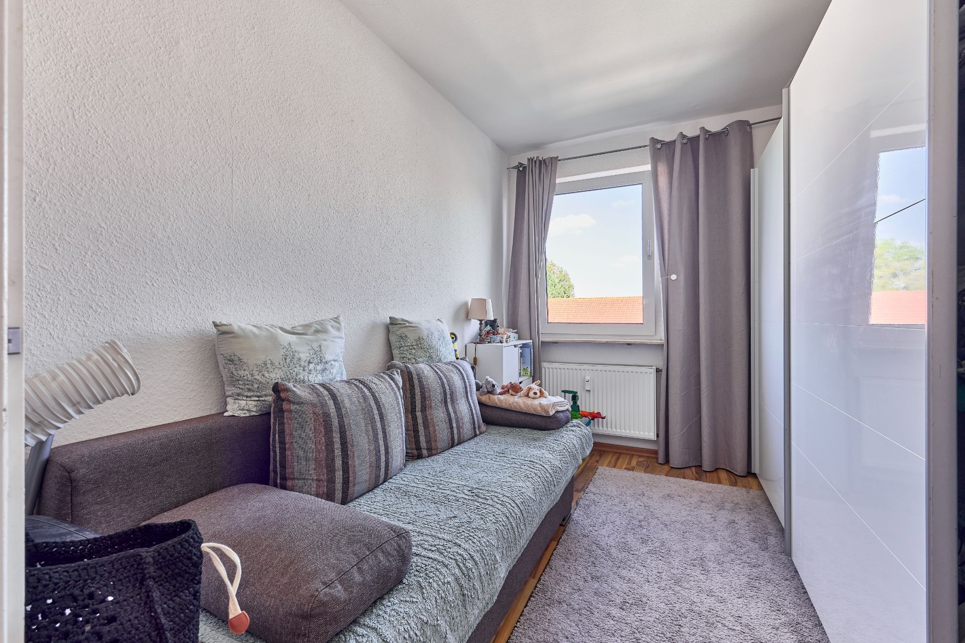 Kleines Zimmer mit grauem Sofa, großem weißen Schrank und Fenster mit grauen Vorhängen.