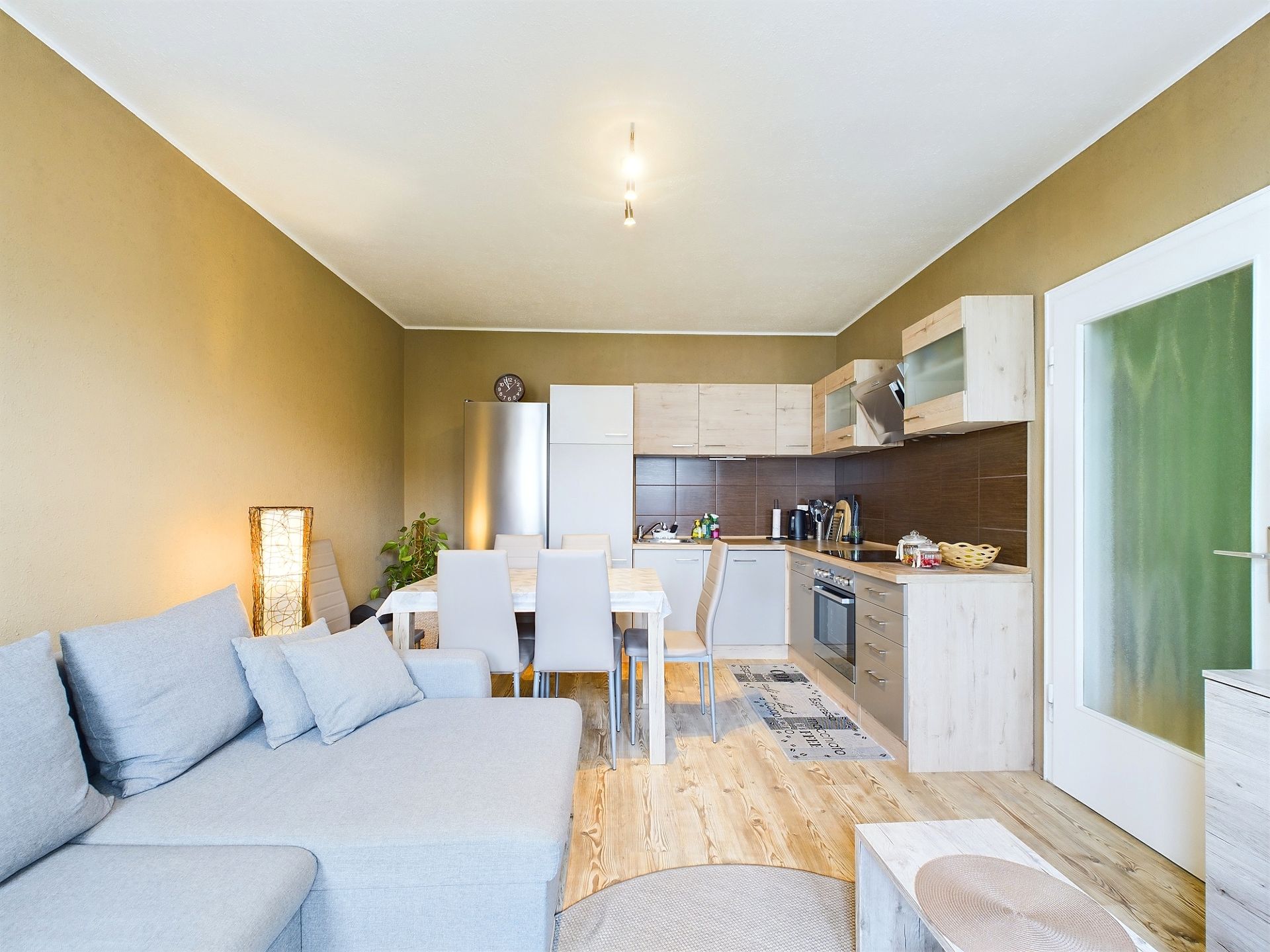 Helles Wohnzimmer mit grauem Sofa, Esstisch, Stühlen und offener Holzküche.