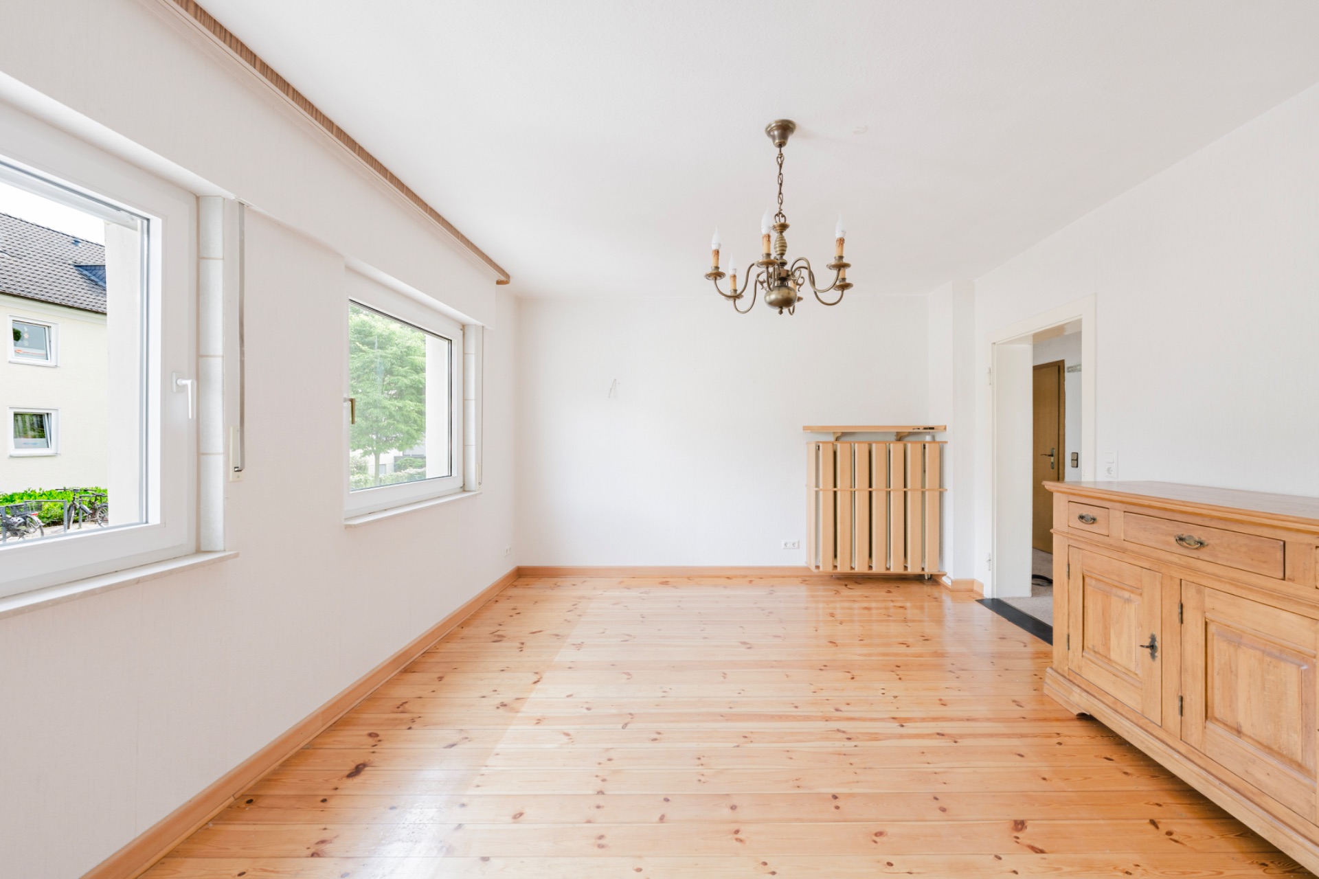 Heller Raum mit Holzboden, zwei Fenstern, Kronleuchter und Holzkommode.