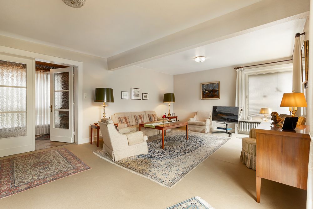 Helles Wohnzimmer mit Teppichen, Sofas, Sessel, Fernseher, Lampen und großen Fenstern.
