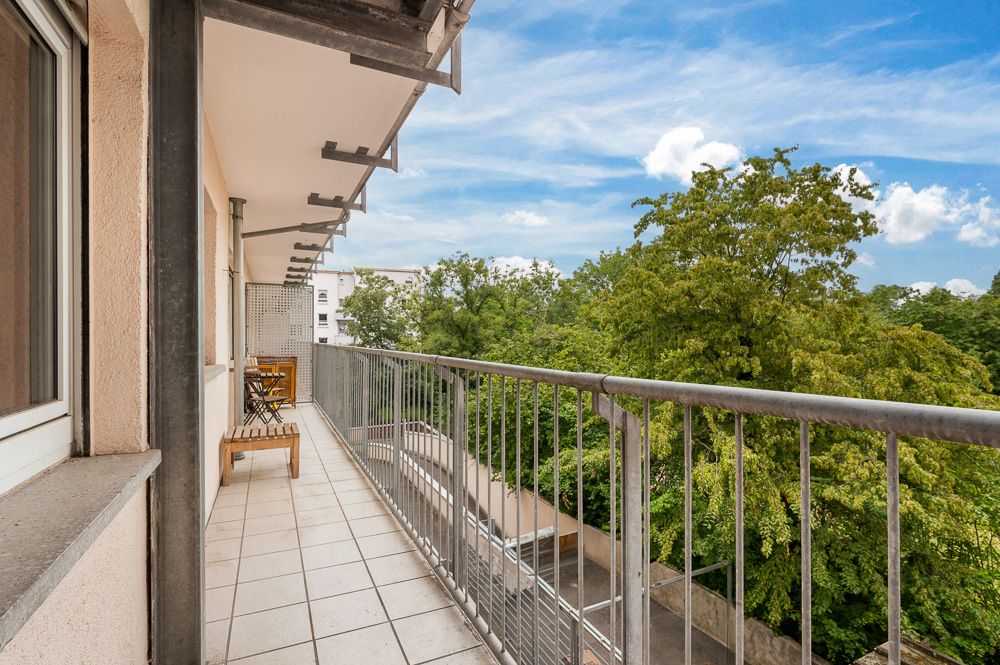 Langer Balkon mit Geländer, Blick auf Bäume, Bank und Stuhl auf den Fliesen.