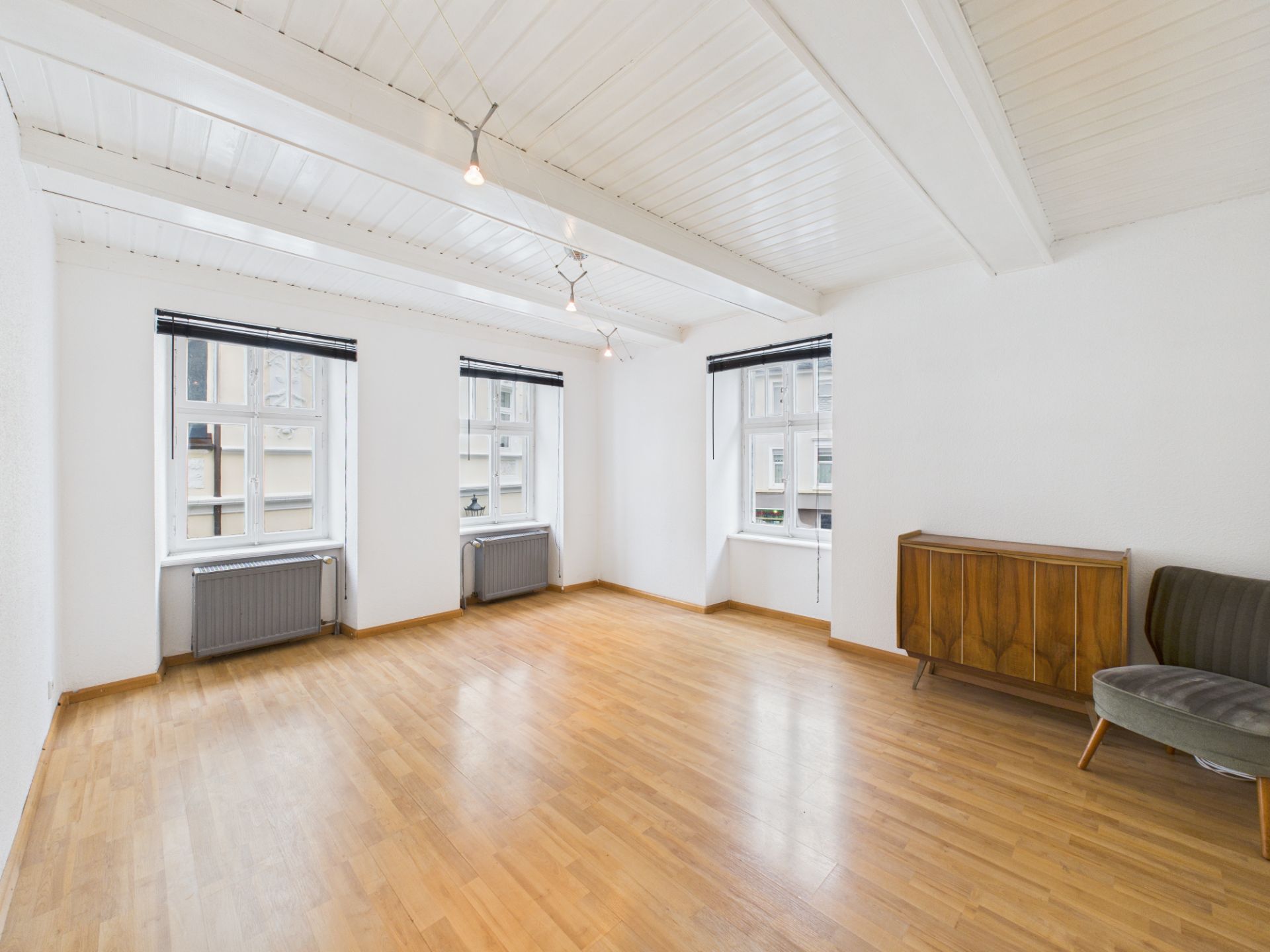 Helles Wohnzimmer mit Holzboden, drei Fenstern, einem Heizkörper, einem Sideboard und einem Sessel.