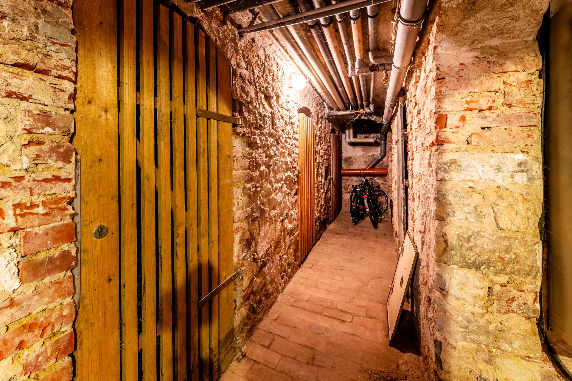 Beleuchteter Kellerflur mit Backsteinwänden, Holztüren, Rohren an der Decke und zwei Fahrrädern am Ende.