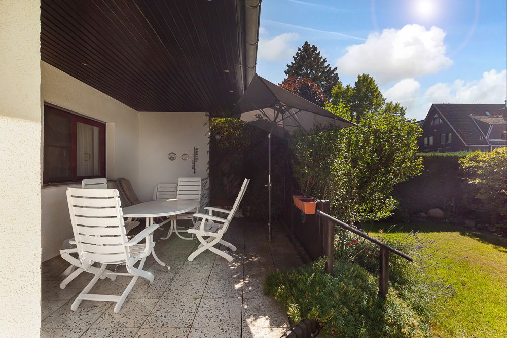 Überdachte Terrasse mit weißen Gartenmöbeln, Sonnenschirm und Blick auf einen grünen Garten.