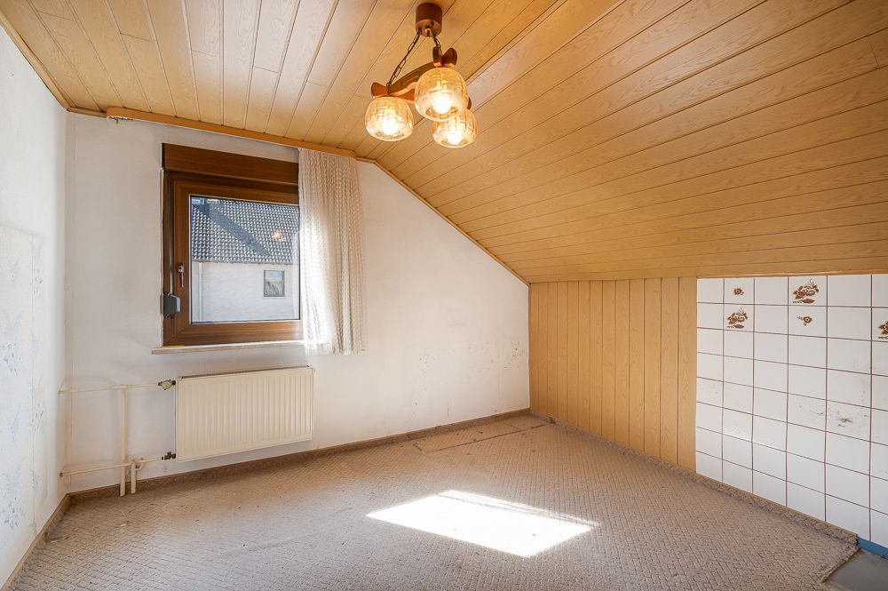 Kleines Zimmer mit Dachschräge, Teppichboden, Fenster, Heizung und Holzdecke.