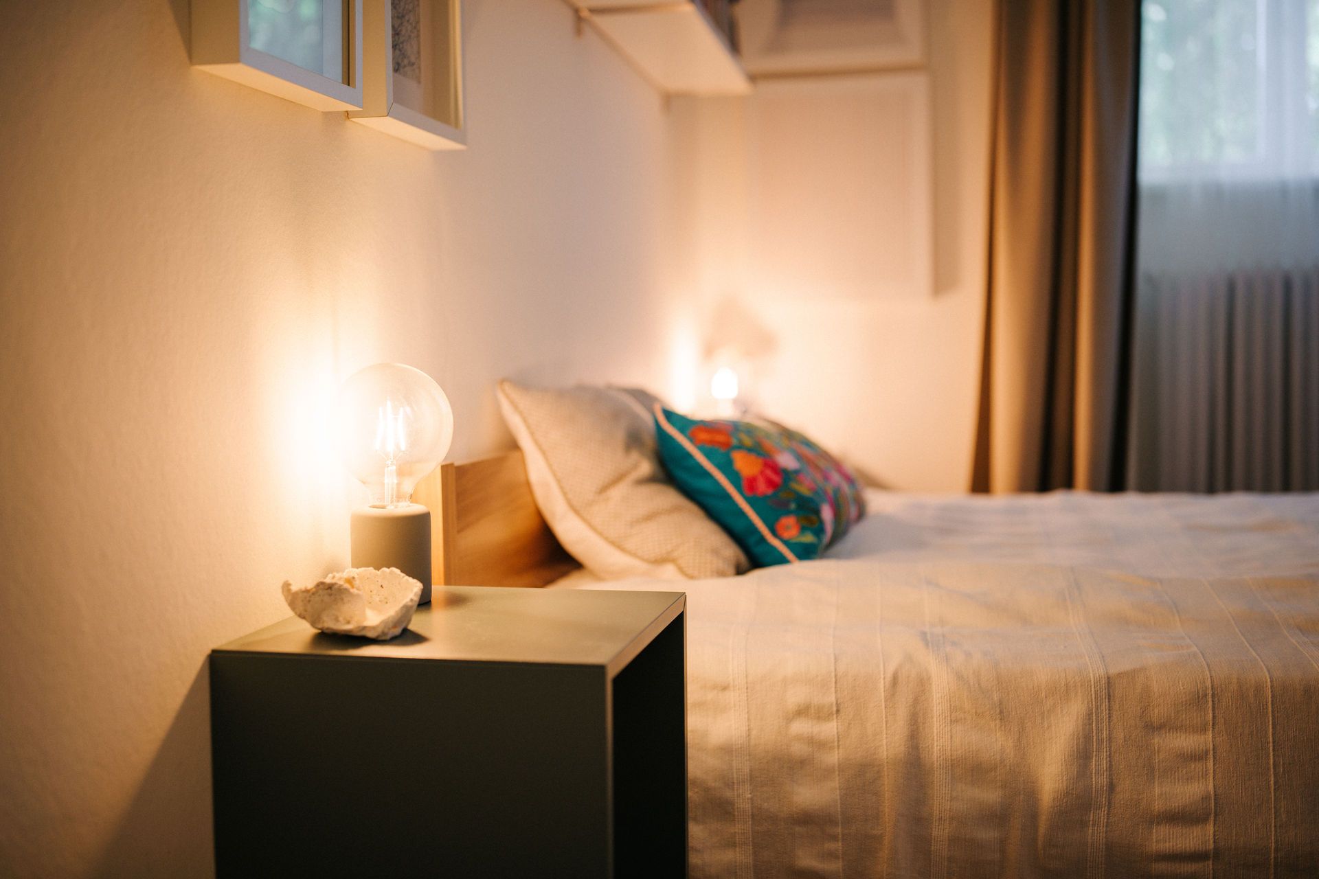 Gemütliches Schlafzimmer mit Nachttisch, Lampe, Bett und buntem Kissen.