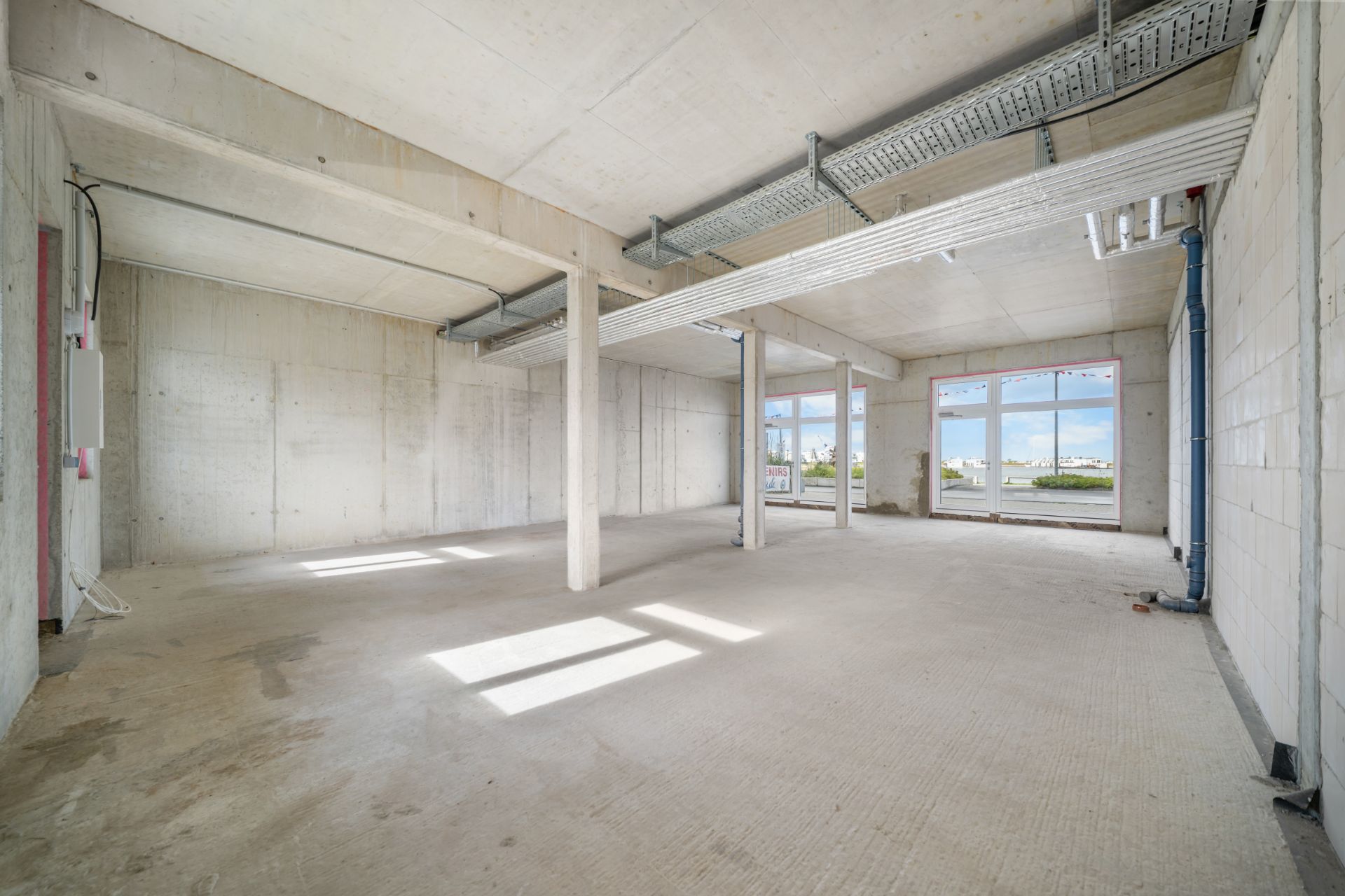 Leerer Rohbau-Raum mit Betonwänden, großen Fenstern und sichtbaren Leitungen an der Decke.