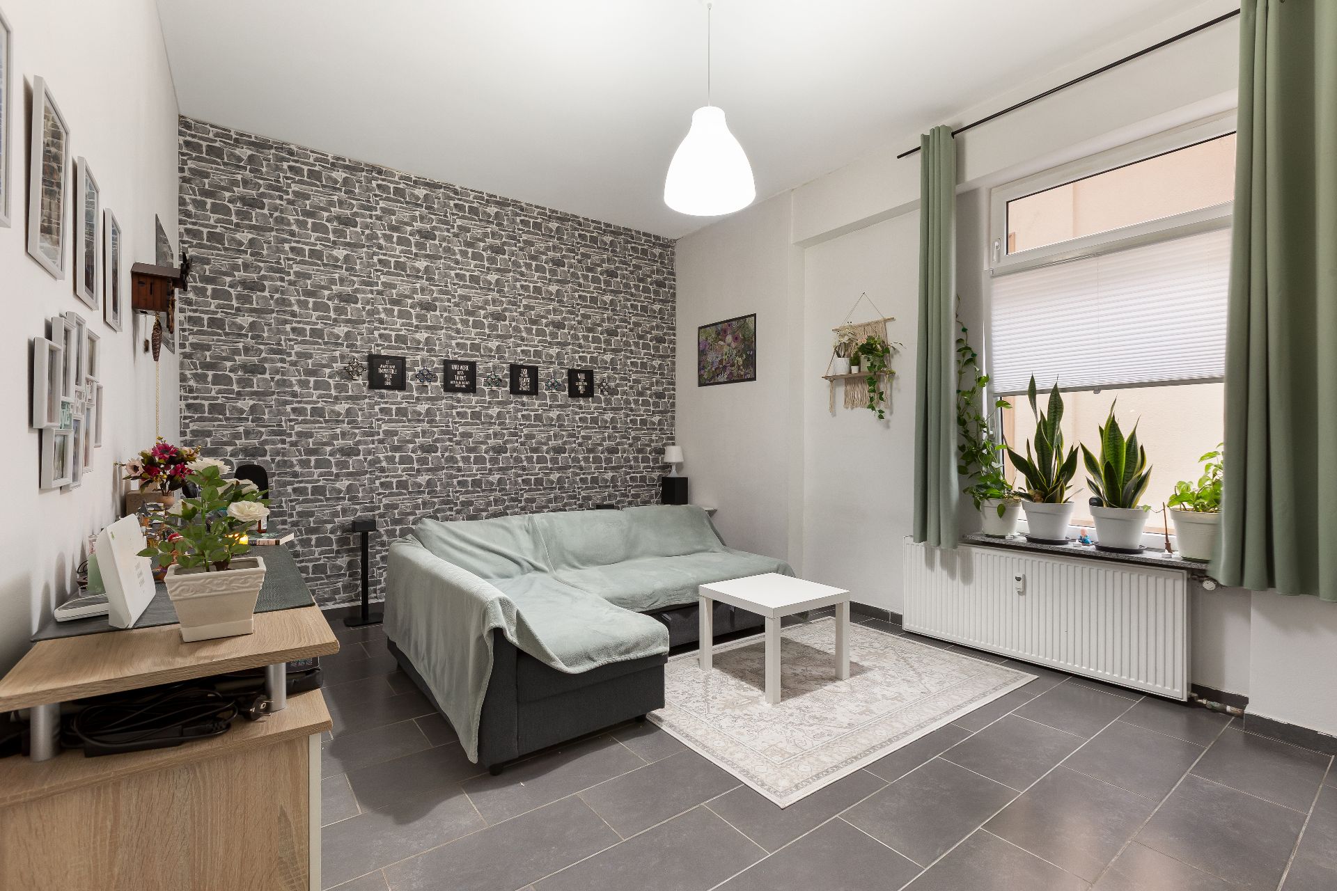 Wohnzimmer mit grauem Ecksofa, weißem Tisch, Steinmusterwand und vielen Pflanzen am Fenster.