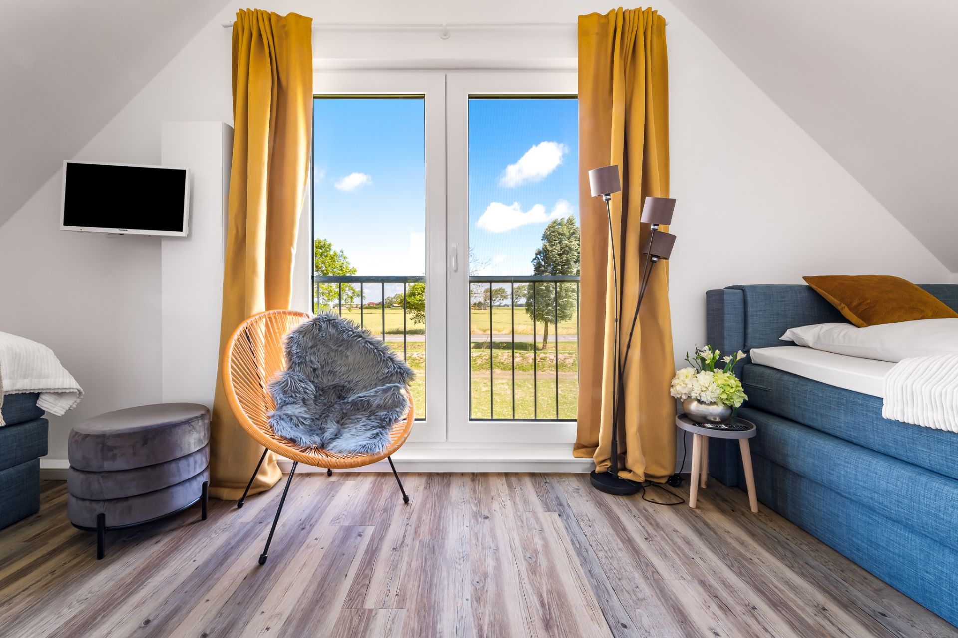 Heller Raum mit Bett, Sessel, Fernseher und großen Fenstern mit gelben Vorhängen.