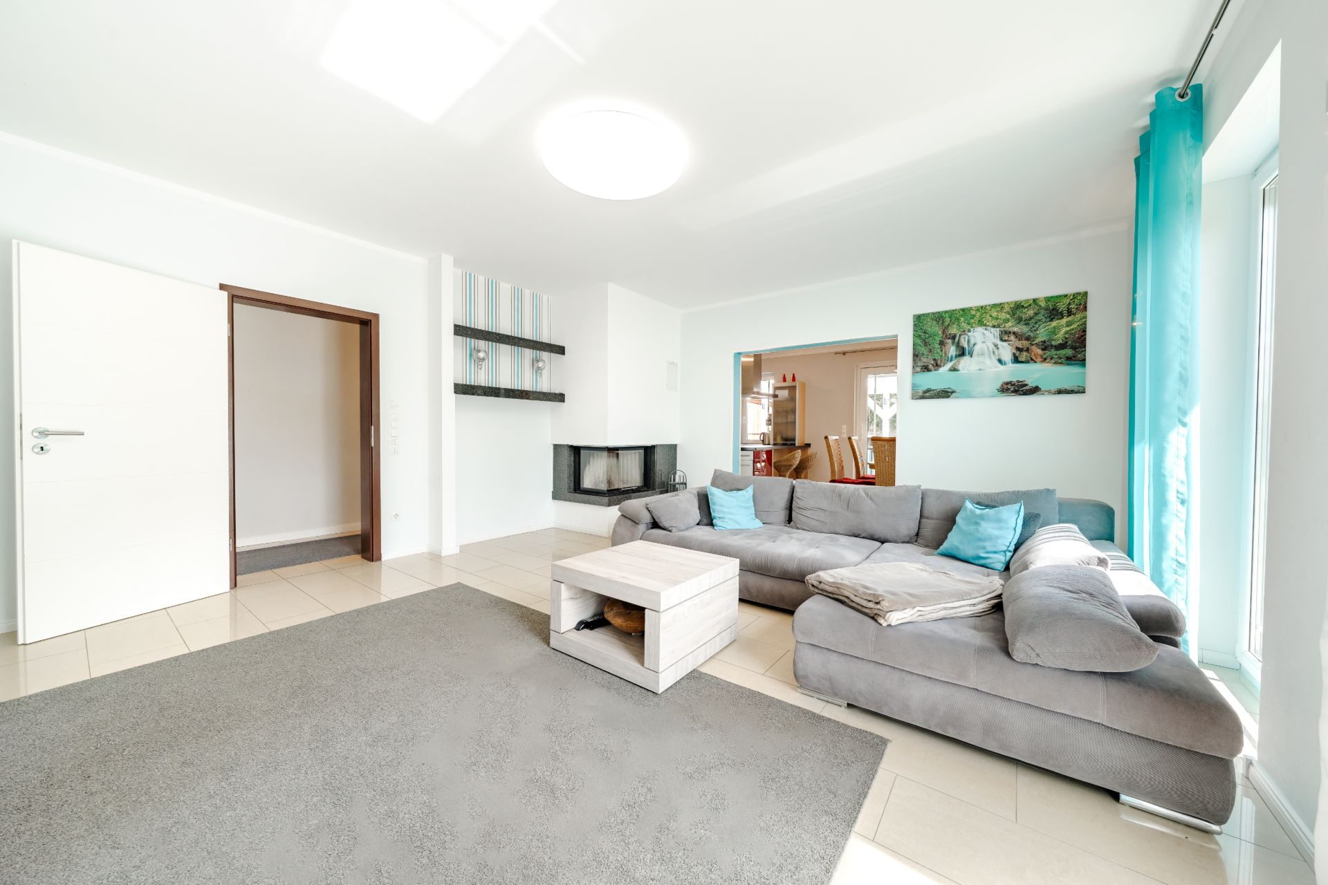 Helles Wohnzimmer mit grauem Sofa, Couchtisch, Teppich und Wandbild, Fenster mit blauen Vorhängen.