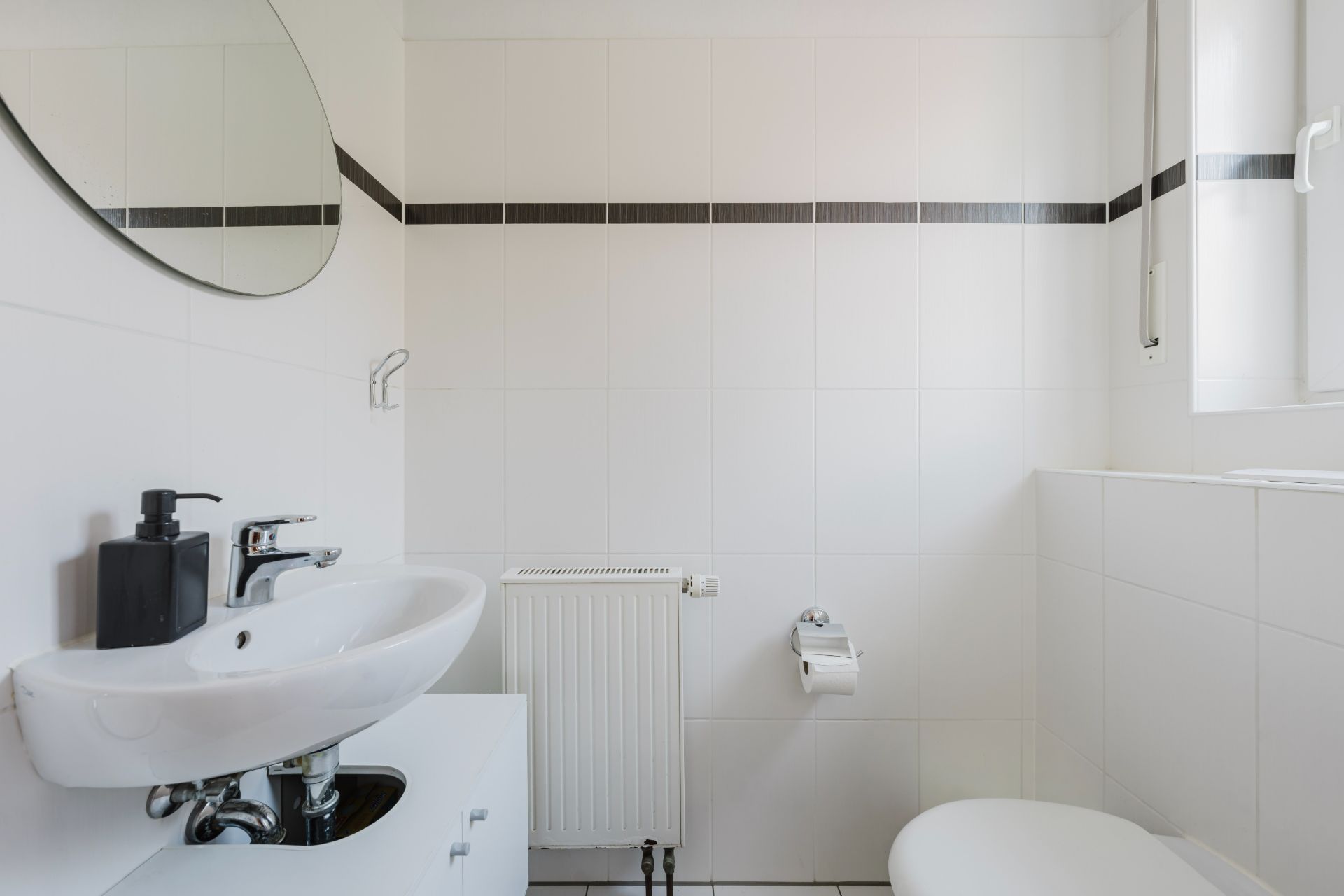 Modernes, weiß gefliestes Bad mit Waschbecken, Spiegel, WC und Fenster.