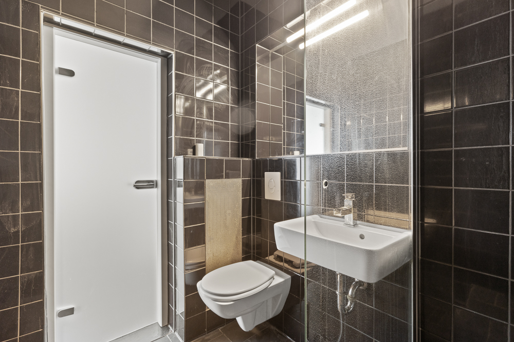 Modernes Bad mit dunklen Fliesen, Wand-WC, Waschbecken und Glasdusche.
