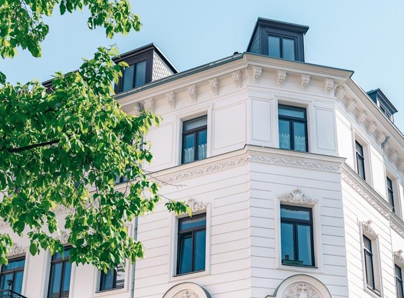 Fassade eines weißen Wohnhauses mit großen Fenstern und Dachgauben, davor ein grüner Baum.