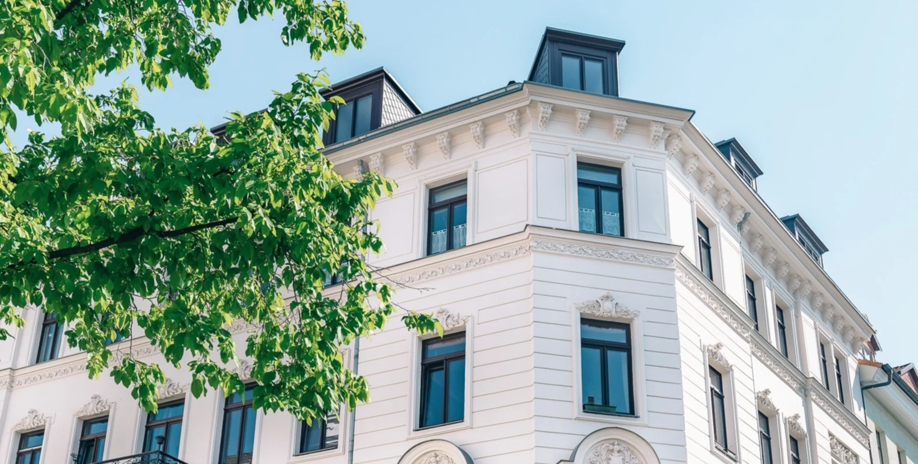 Fassade eines weißen Wohnhauses mit großen Fenstern und Dachgauben, davor ein grüner Baum.