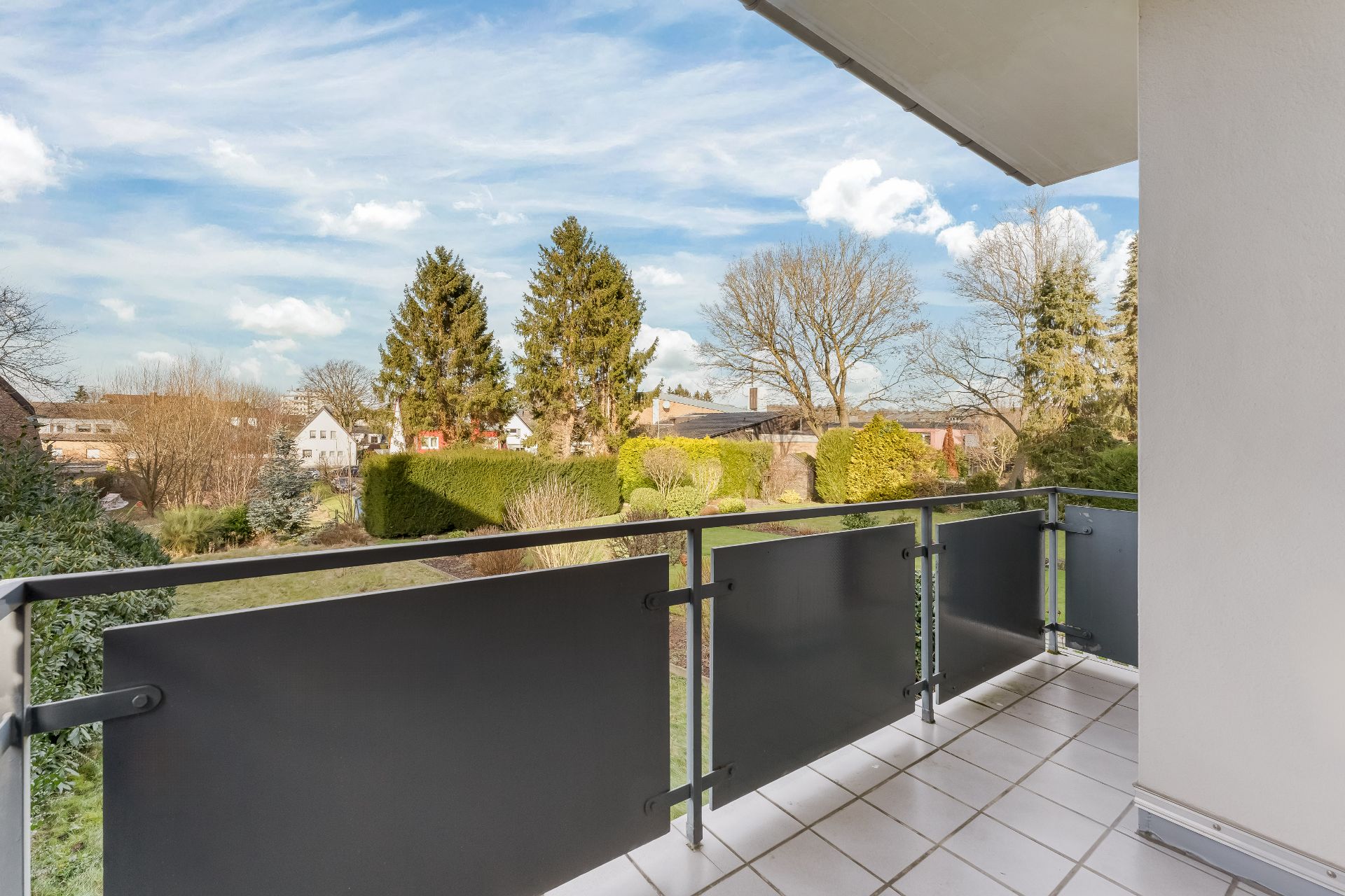 Balkon mit Blick auf einen grünen Garten und Bäume, darunter einige Wohnhäuser im Hintergrund.